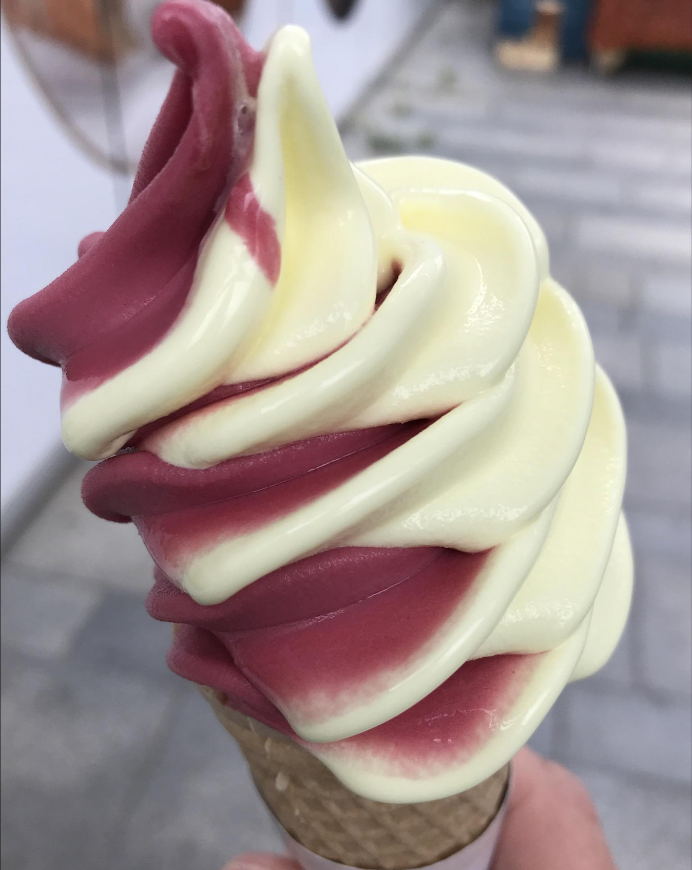 A close-up of a hand holding a swirled ice cream cone with yellow and pink flavors, set against a blurred outdoor background with gray pavement.