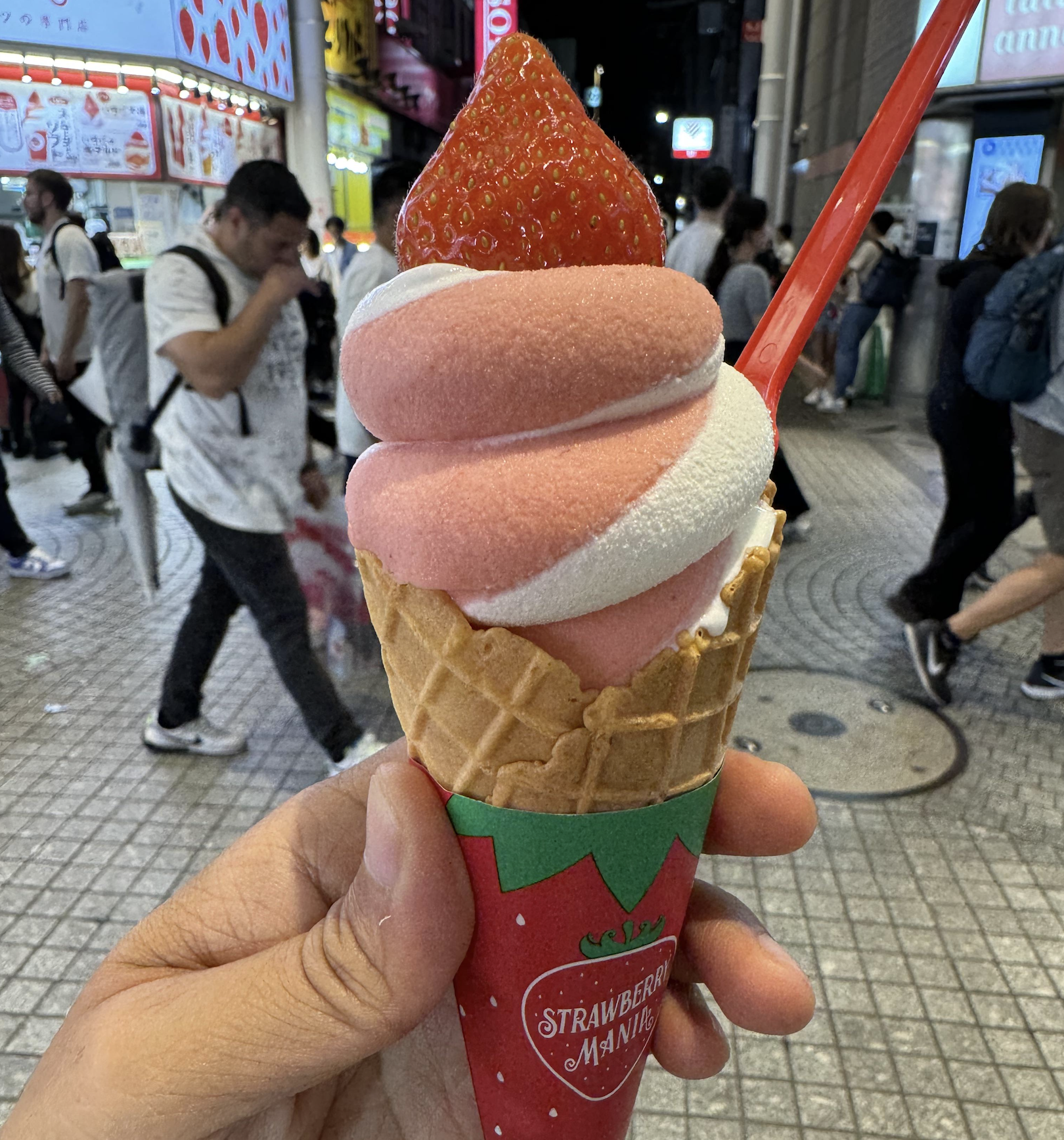 A hand holds a strawberry and vanilla swirl ice cream cone with a fresh strawberry on top. People walk by on a busy, brightly lit city street in the background.