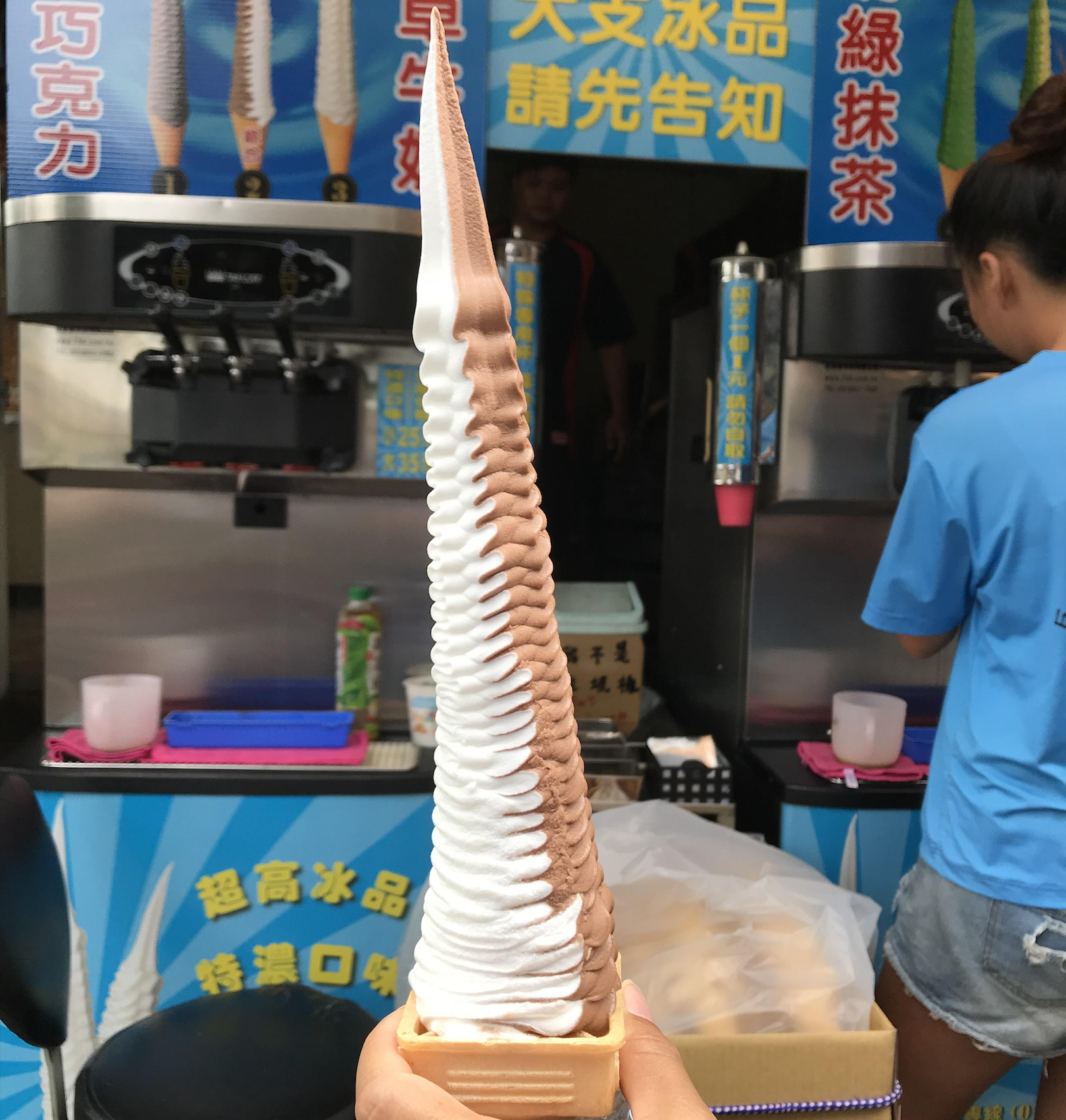 A very tall, swirled chocolate and vanilla soft-serve ice cream cone is held up in front of a colorful ice cream stand, with a person in a blue shirt standing nearby.