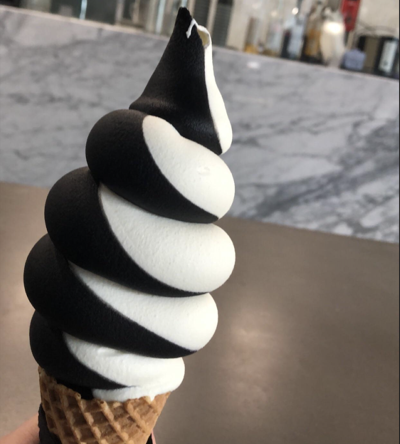 A hand holds a waffle cone with swirled black and white soft serve ice cream. A blurred marble counter and various items are in the background.