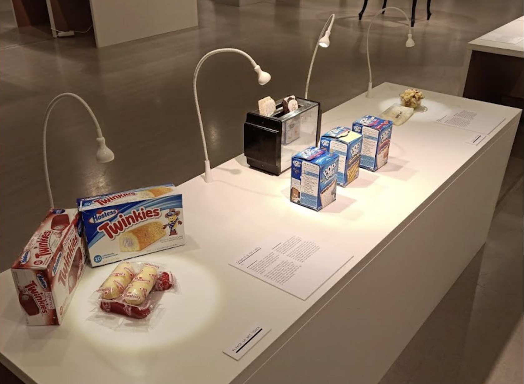 A display case featuring boxes of Twinkies, several cartons of milk, and a black toaster, each item illuminated by a small white lamp on a white platform in a gallery setting.