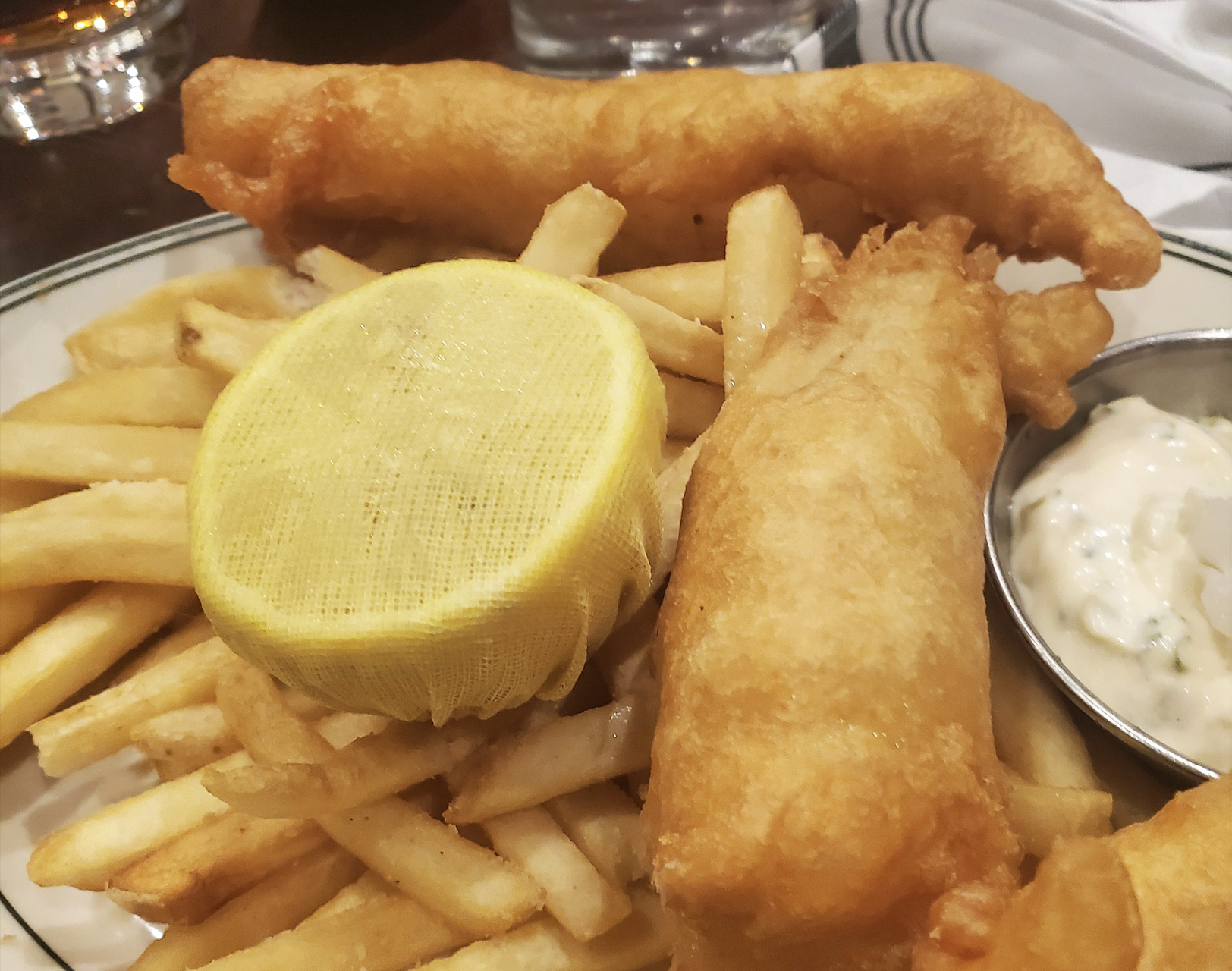 A plate of fish and chips with two battered fish fillets, French fries, a lemon half covered with mesh, and a small cup of tartar sauce on the side.