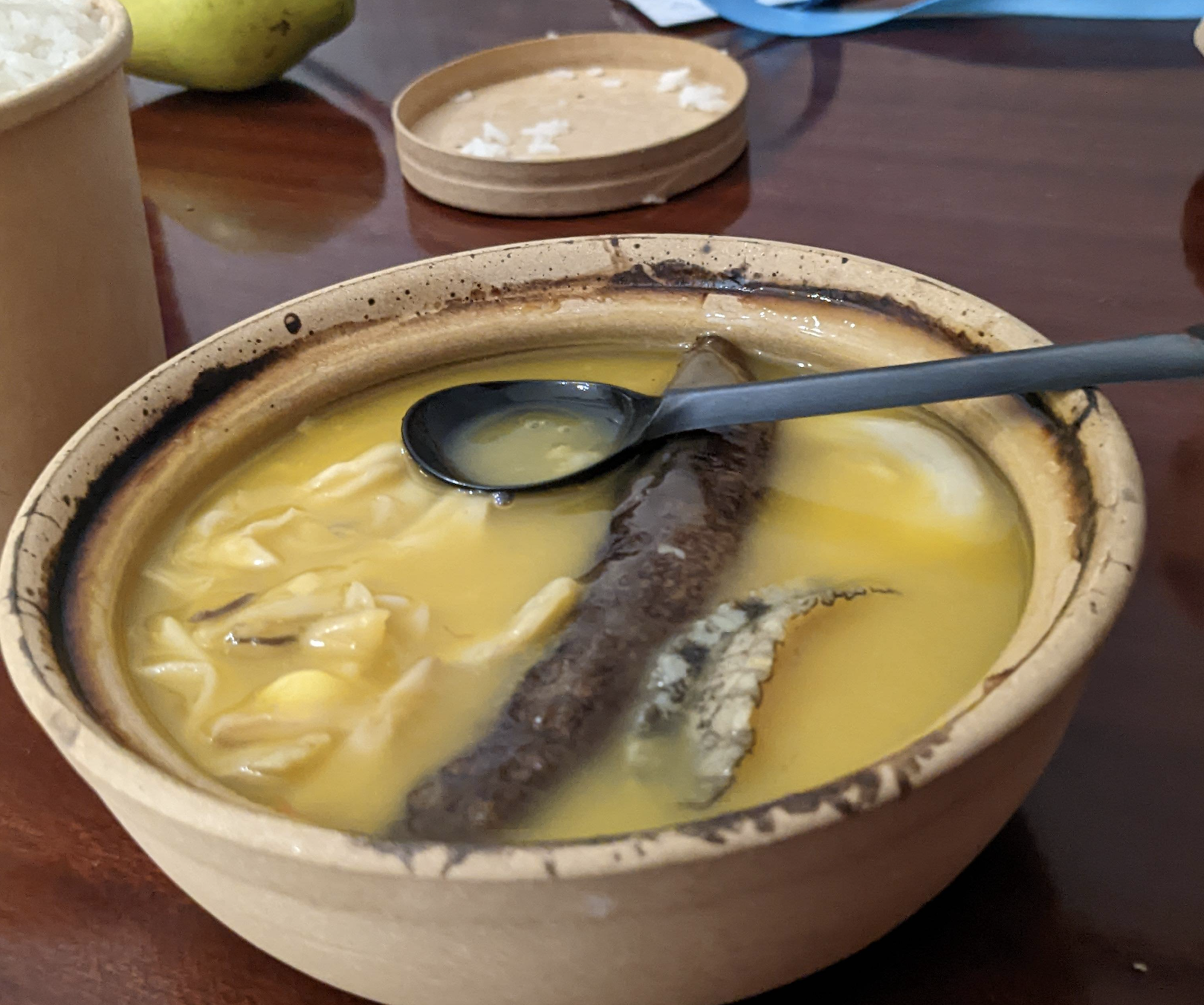 A bowl of yellow soup with a dark sausage, cabbage, and a black spoon on a wooden table, with rice and a pear in the background.