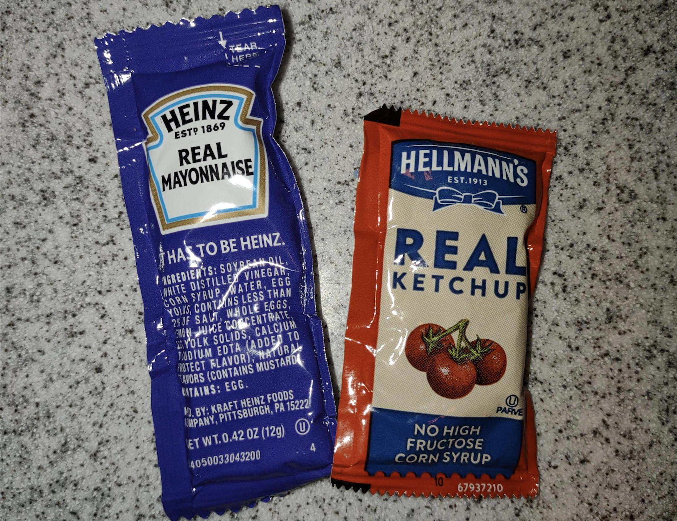 Two condiment packets on a speckled countertop: a blue Heinz Real Mayonnaise packet and a red Hellmann’s Real Ketchup packet with tomatoes pictured on it.