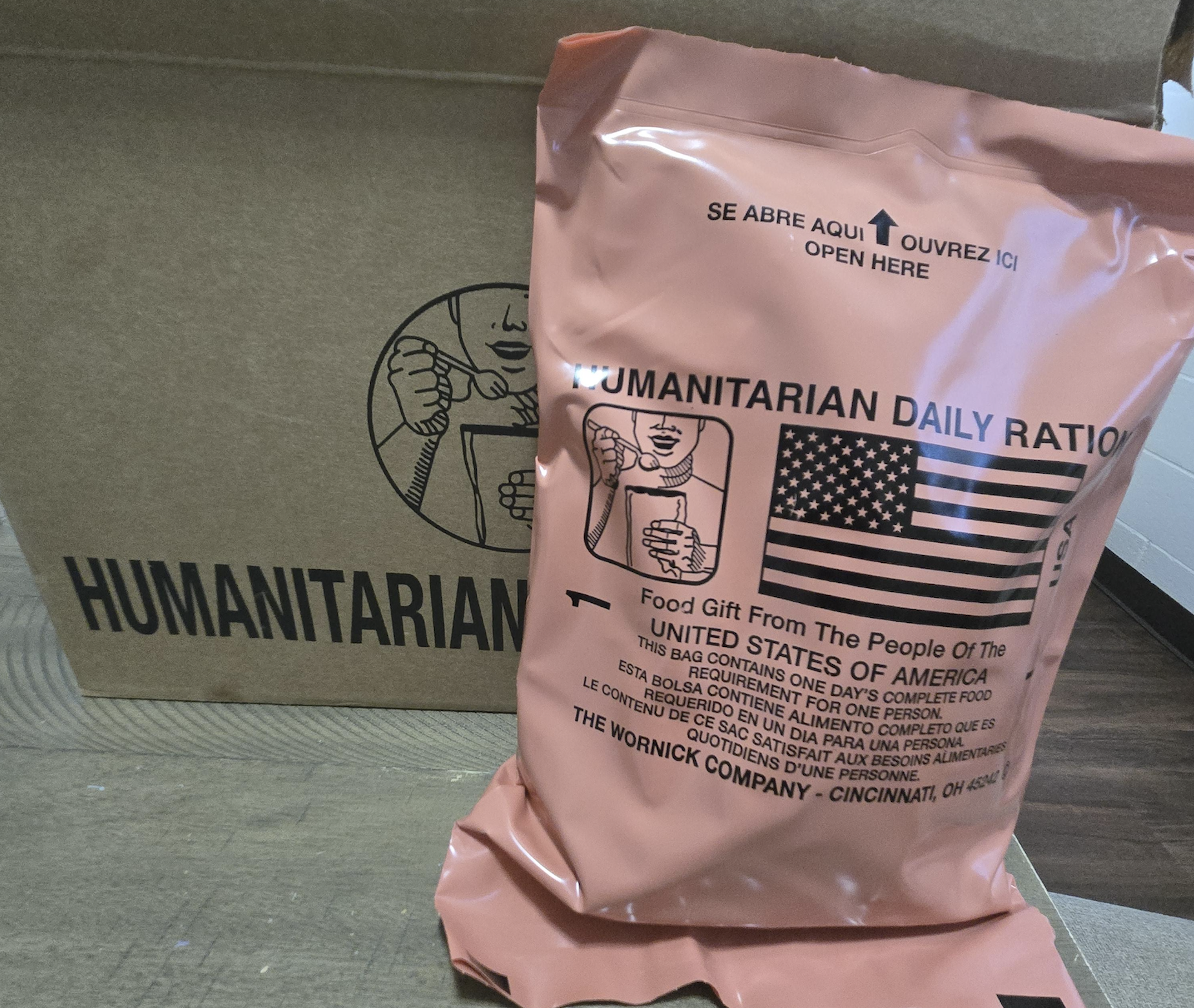 A cardboard box and a sealed orange Humanitarian Daily Ration food pouch labeled with the U.S. flag and instructions for opening, meant for emergency food aid.