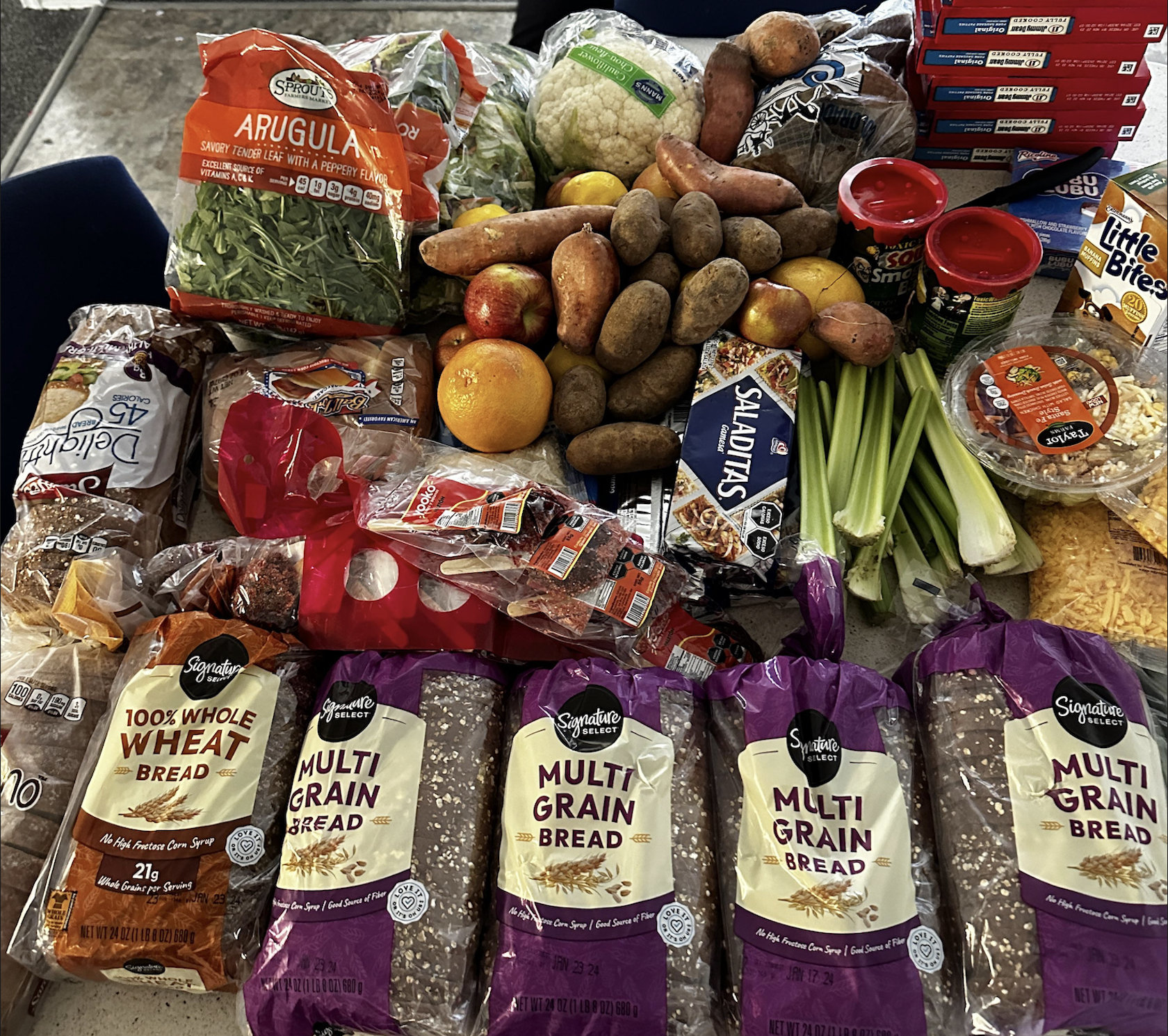 A variety of groceries on a table, including multigrain bread, wheat bread, lettuce, potatoes, apples, celery, cheese, pasta, snack cakes, and other packaged food items.