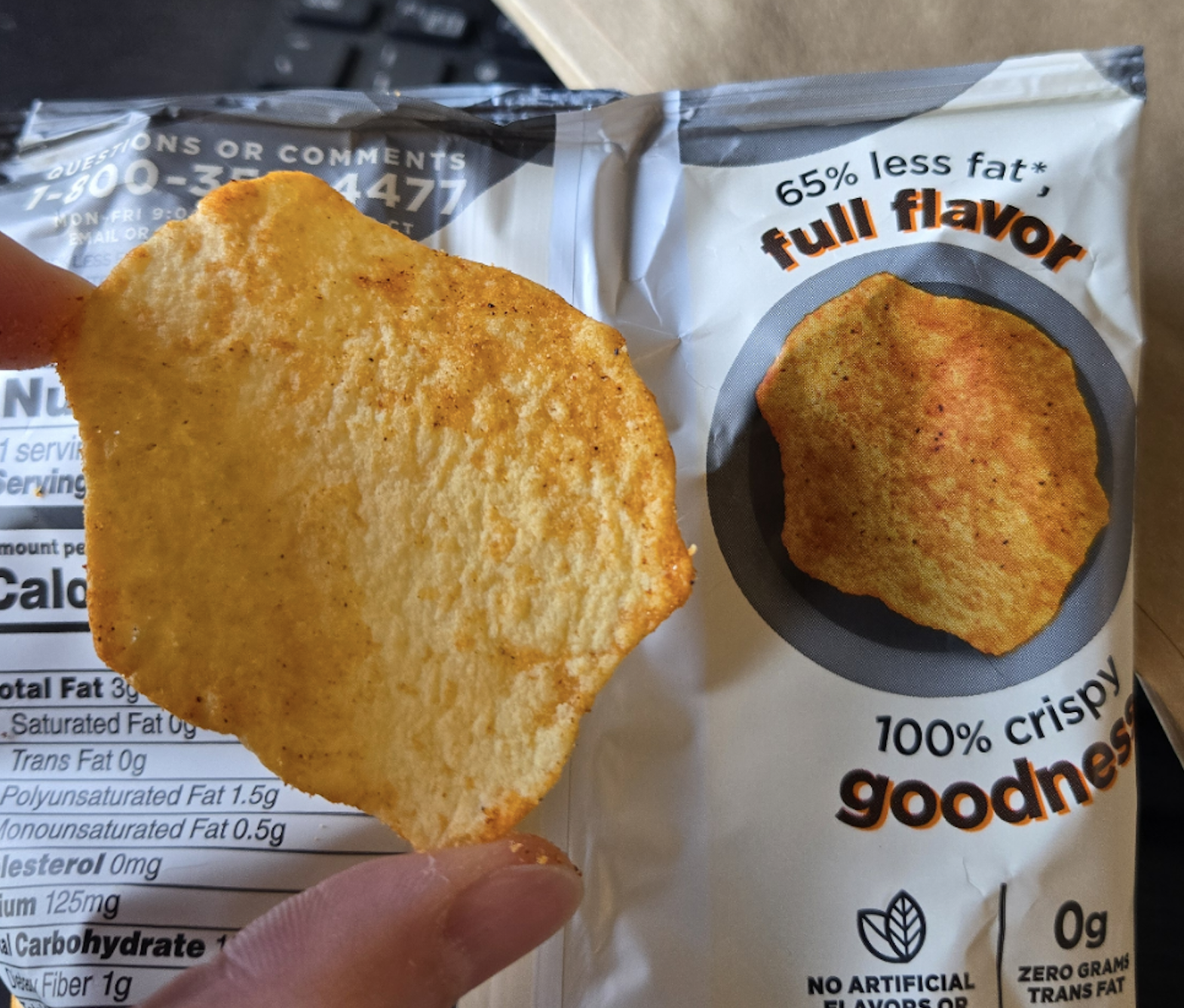 A hand holds a round, golden potato chip next to a snack bag showing a similar chip image. The bag advertises “65% less fat,” “full flavor,” and “100% crispy goodness.”