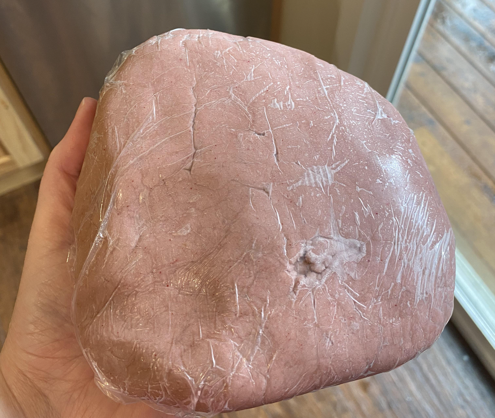 A hand holds a large lump of pinkish dough wrapped in plastic wrap, with a small indentation on the surface. A wooden floor and glass door are visible in the background.
