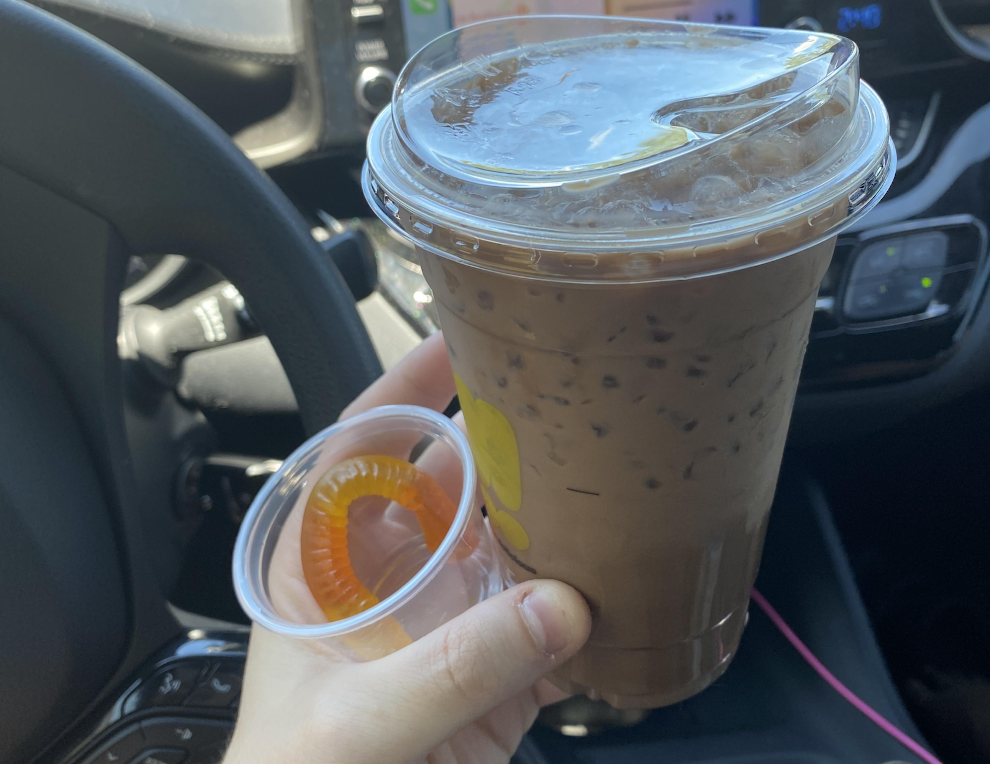 A hand holds an iced chocolate drink in a plastic cup with a domed lid and straw, along with a small plastic cup containing a gummy worm, inside a car.