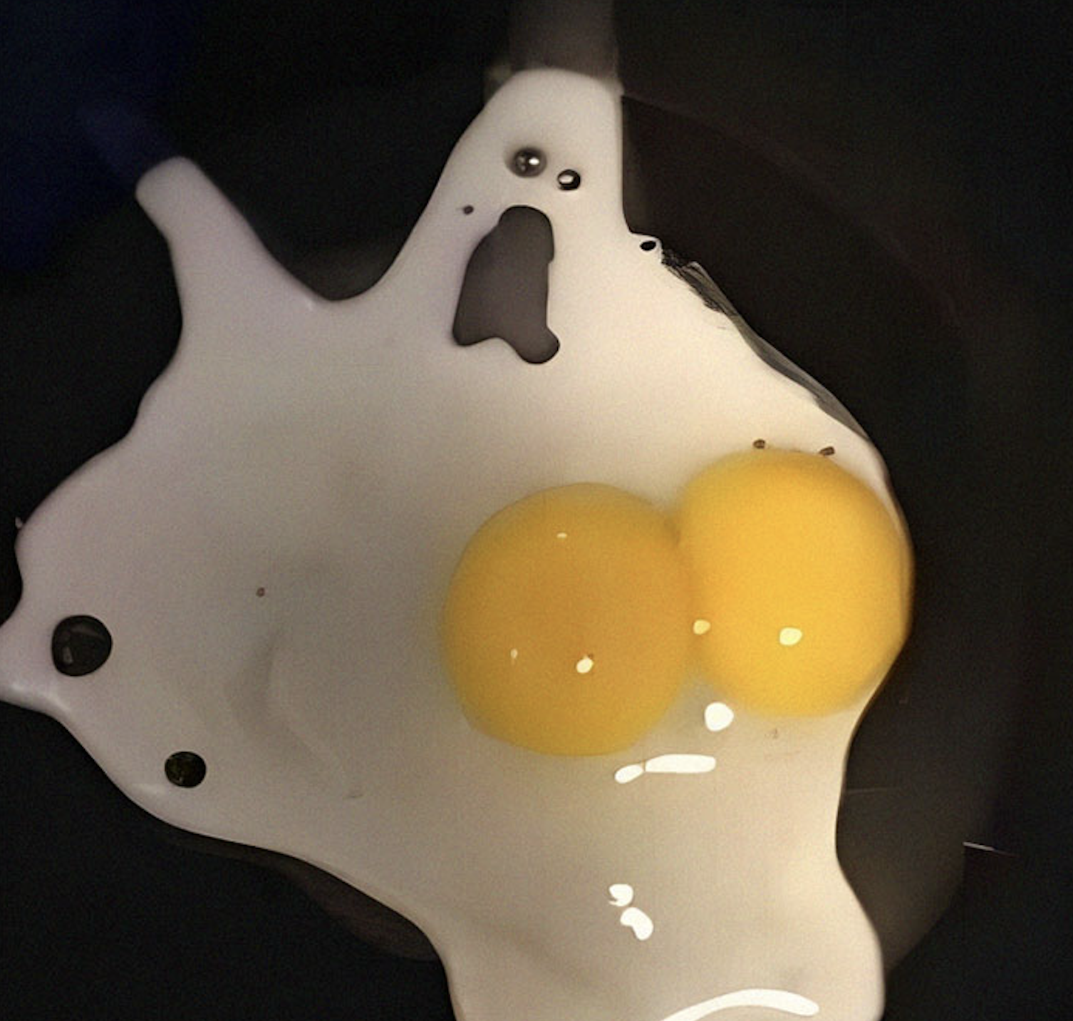 A cracked egg with two yolks sits on a dark surface. The egg white has spread in a shape that resembles a surprised ghost face near the top, with two small dots for eyes and an open mouth.