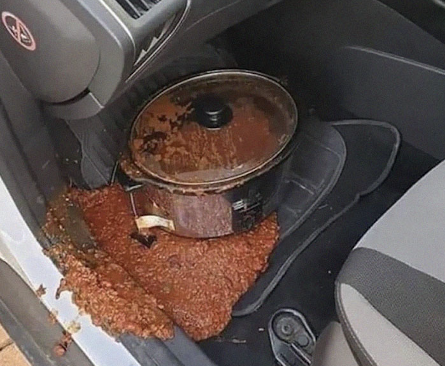 A slow cooker spilled chili all over the front passenger side floor mat of a car, creating a messy scene.