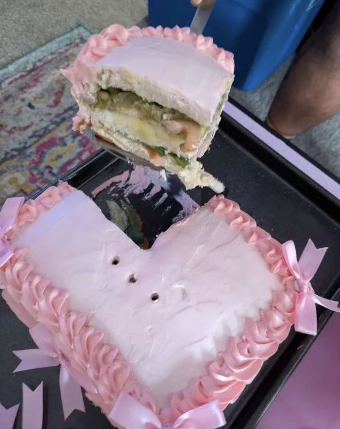 A heart-shaped cake with pink frosting and bows has a slice removed, revealing layers of sandwich fillings like lettuce and tomato inside instead of cake.