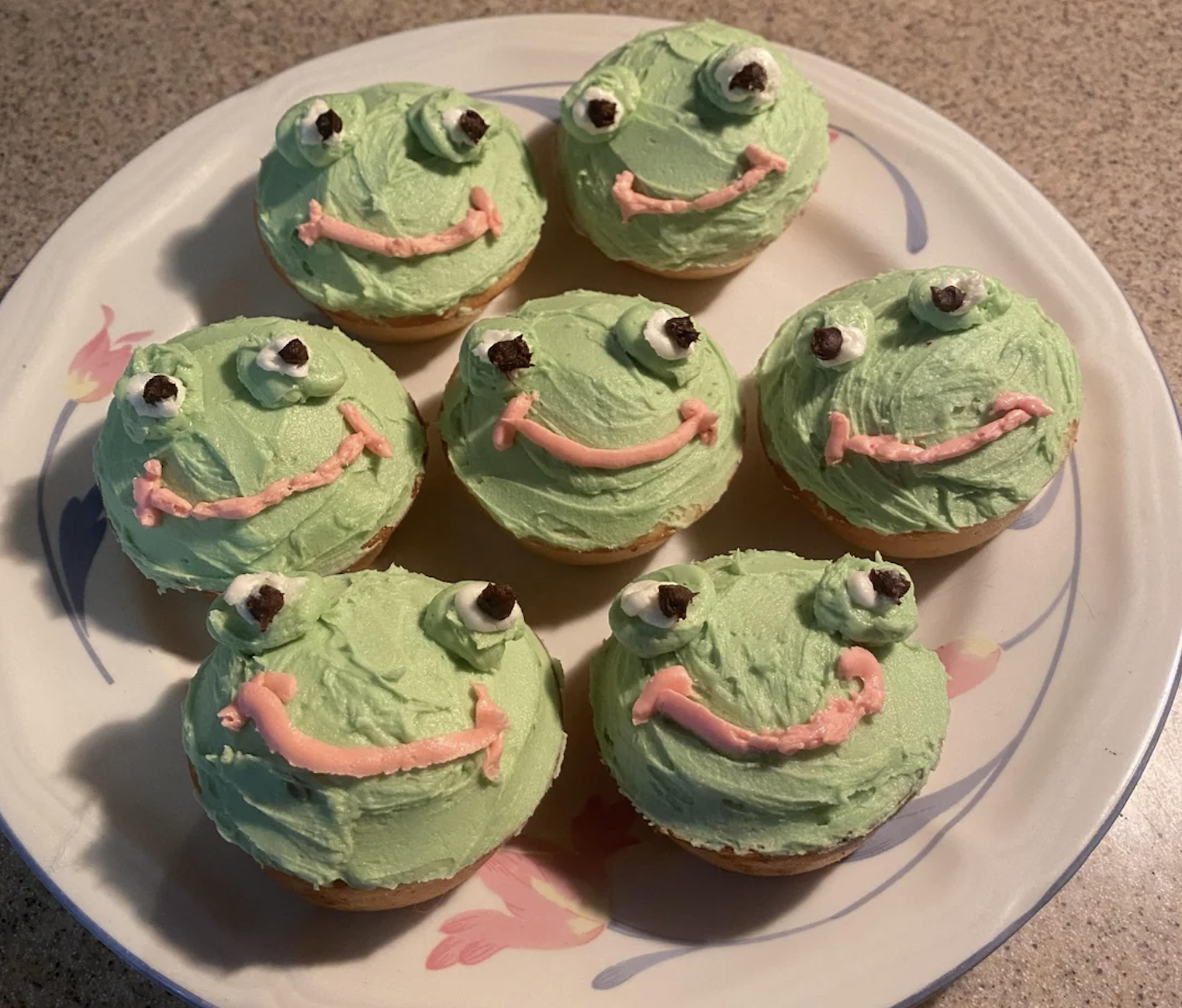 Seven cupcakes decorated with green frosting, pink mouths, and candy eyes to look like smiling cartoon frogs, arranged on a white plate with pink designs on a countertop.