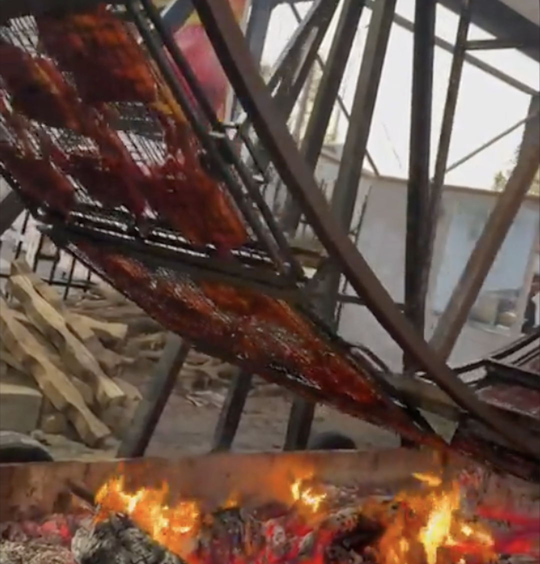 Large metal grill racks rotate above an open fire, roasting slabs of meat. Stacks of wood sit nearby, and metal beams form part of the background structure.