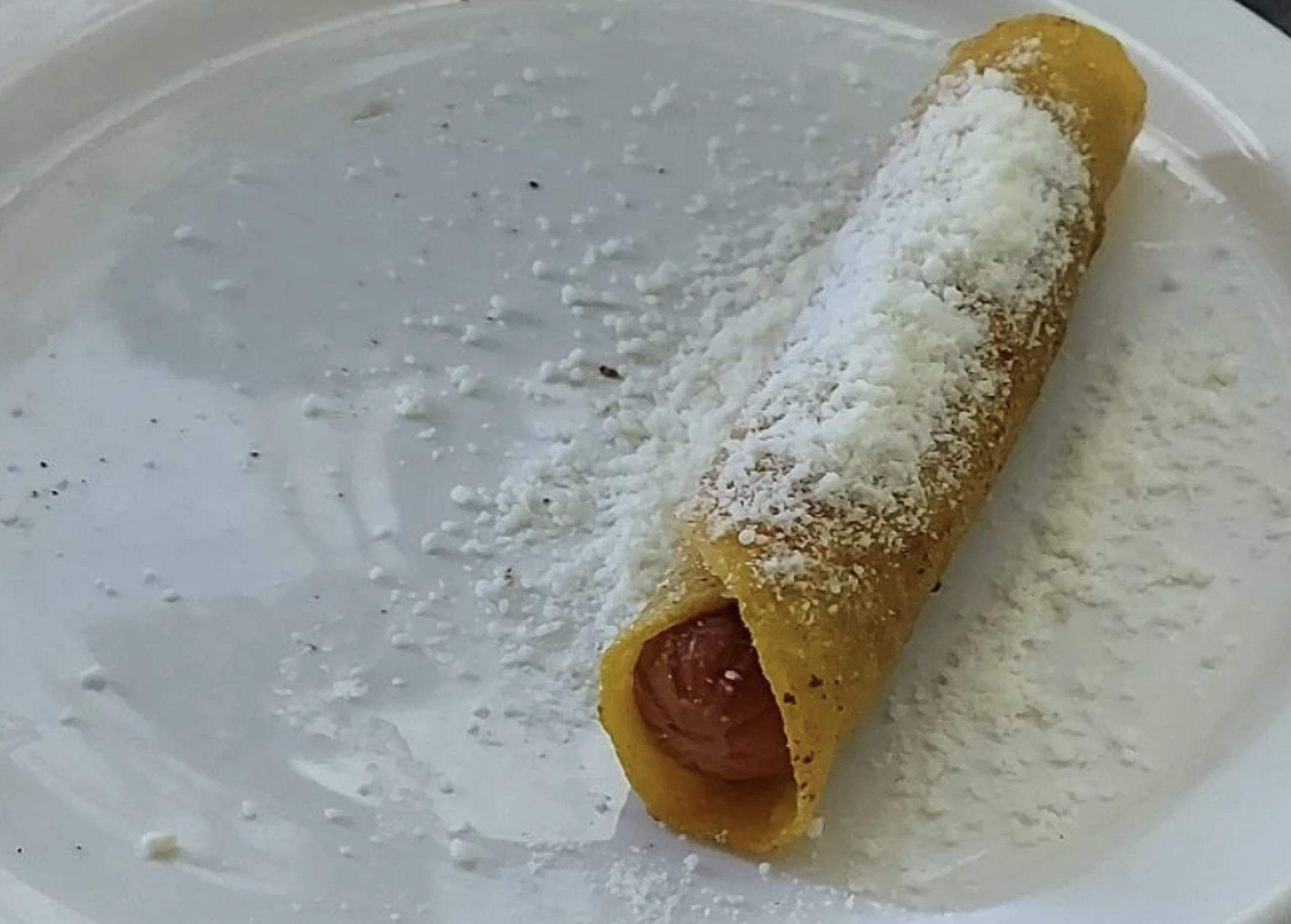 A hot dog wrapped in a corn tortilla, topped with a dusting of shredded white cheese, sits on a white plate with scattered cheese around it.