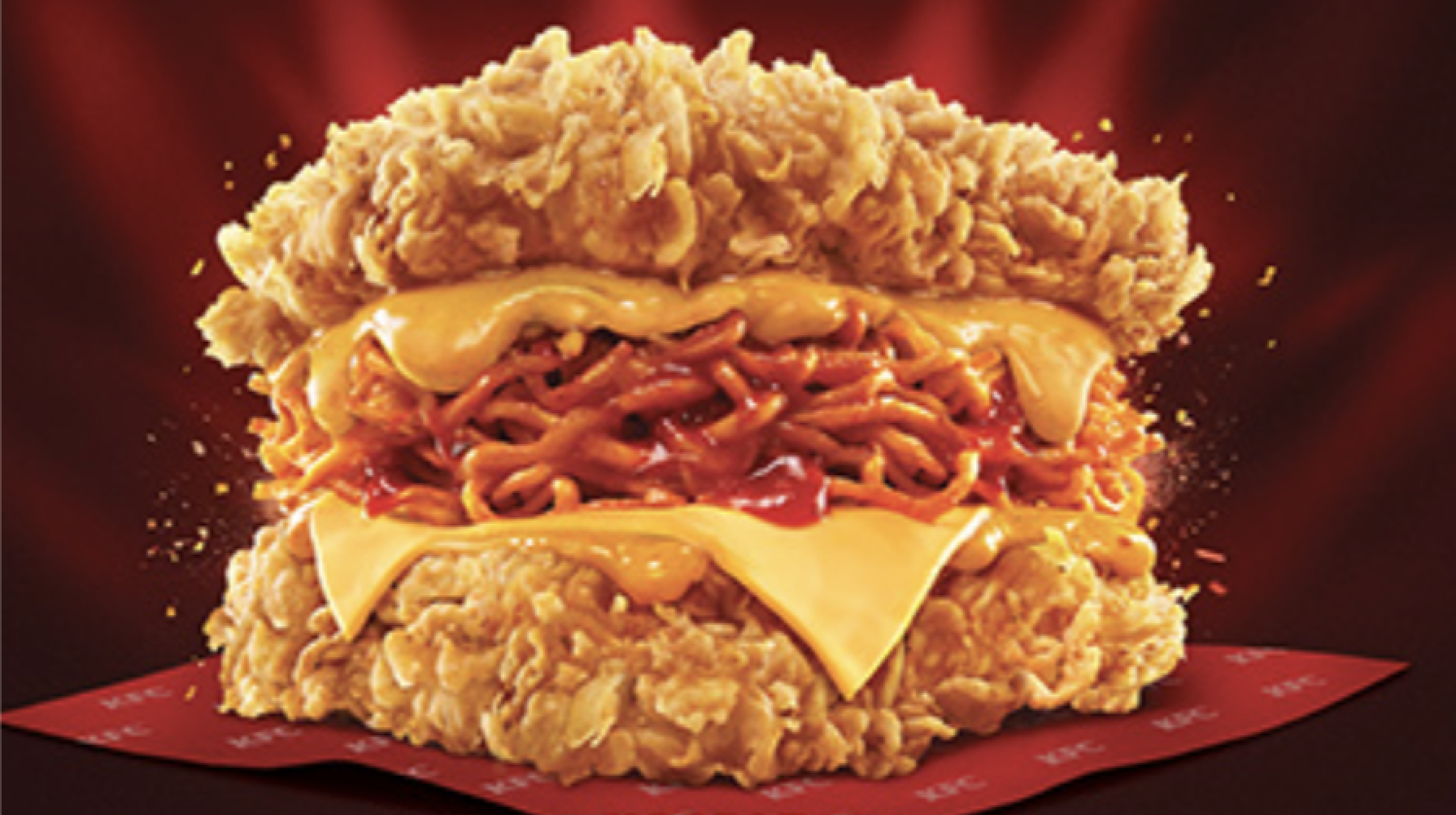 A sandwich with two pieces of fried chicken as buns, filled with noodles, melted cheese, and sauce, on a KFC-branded wrapper with a dark red background.