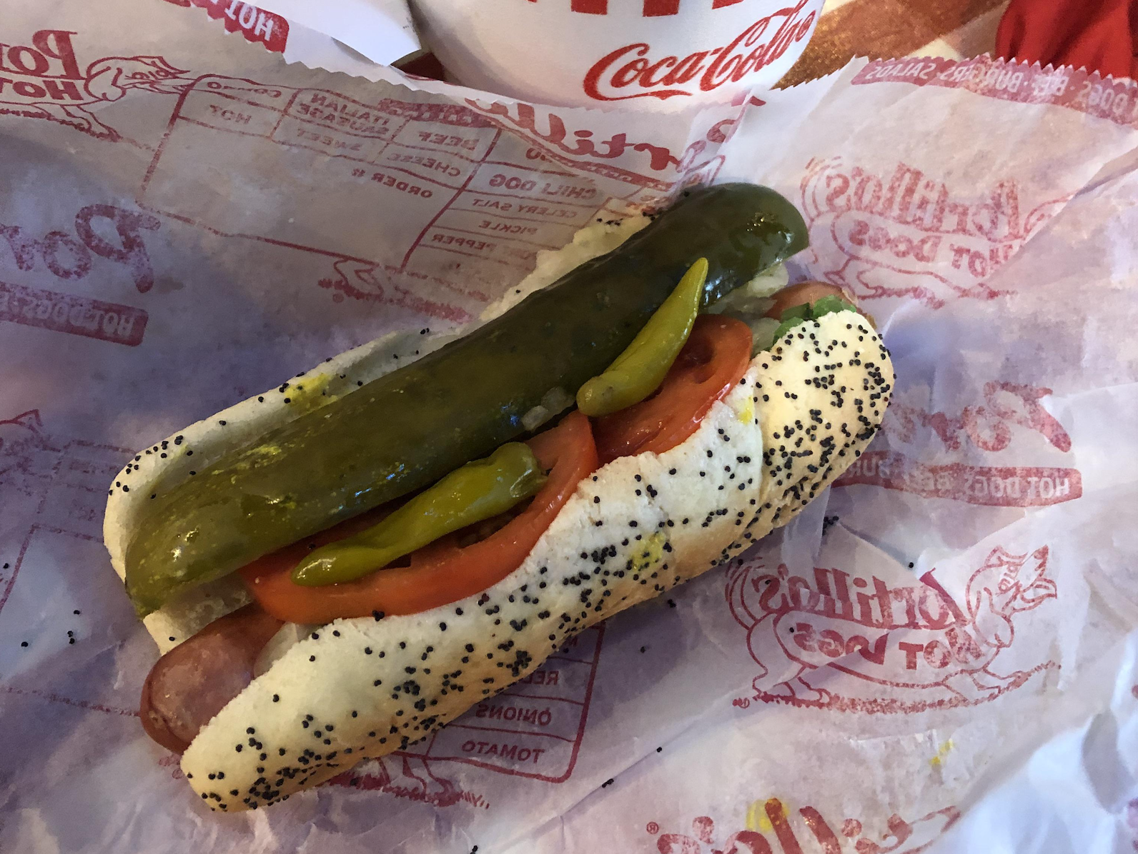 A poppy seed bun hot dog topped with a pickle spear, tomato slices, and sport peppers on paper with a Coca-Cola cup partially visible in the background.