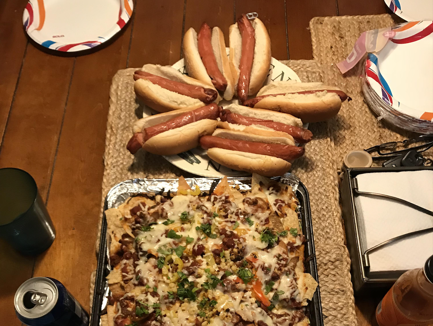 A table with six hot dogs on buns, a tray of loaded nachos with melted cheese, a can of soda, a drink, paper plates, and a napkin holder set on a woven mat.