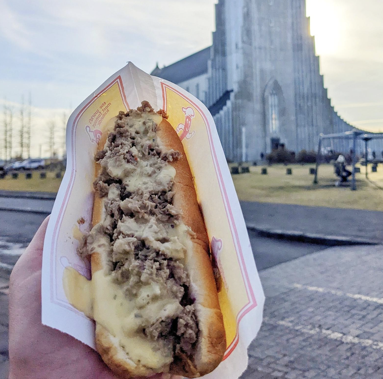 A hand holds a hot dog topped with chopped meat and sauce in front of Hallgrímskirkja church in Reykjavik, Iceland, on a sunny day.