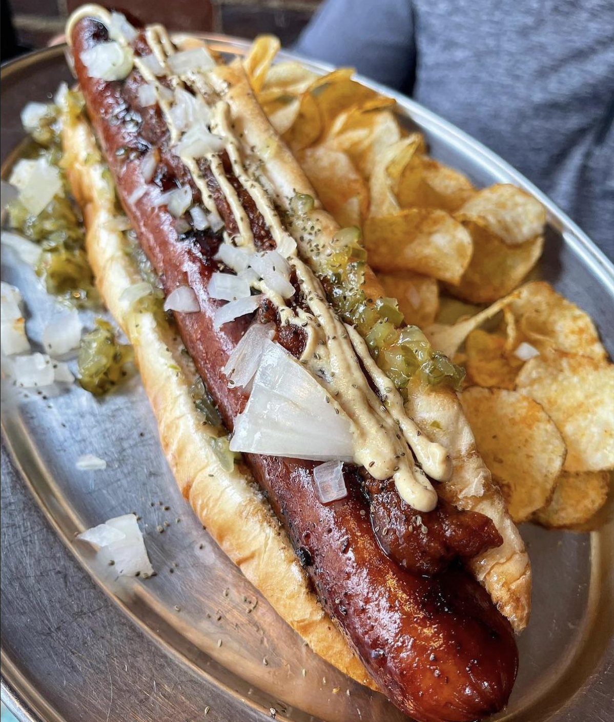 A grilled hot dog in a bun topped with chopped onions, relish, and mustard, served on a metal tray with a side of potato chips.