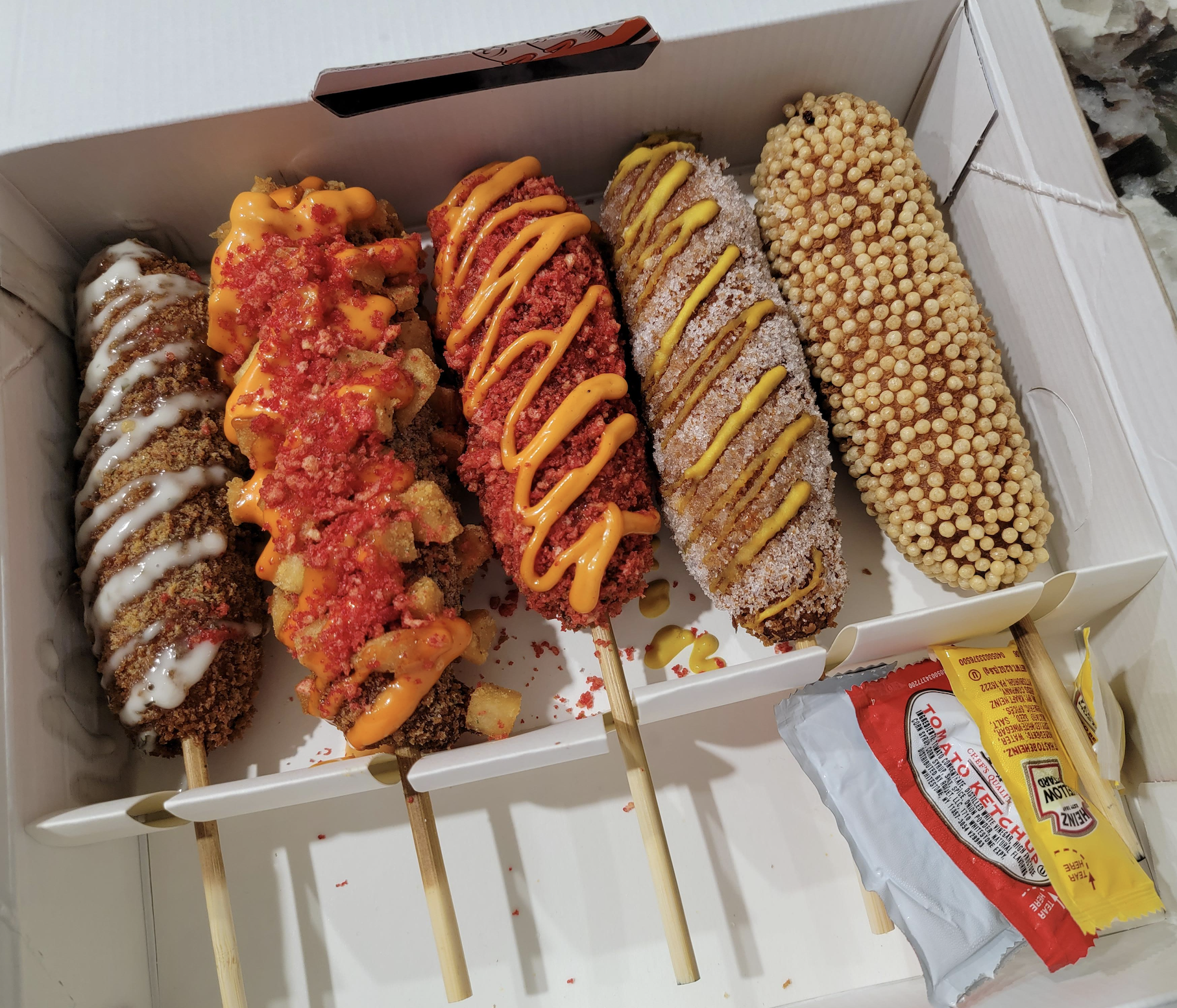 Five Korean corn dogs with various toppings, including cheese, sauces, and crumbs, are arranged in a white box. Condiments like ketchup and mustard packets are placed on the side.