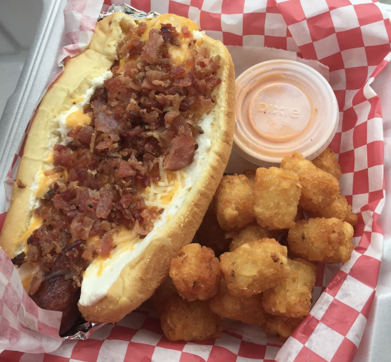 A hot dog topped with shredded cheese, bacon bits, and sauce sits in a red checkered basket next to crispy tater tots and a small cup of orange dipping sauce.