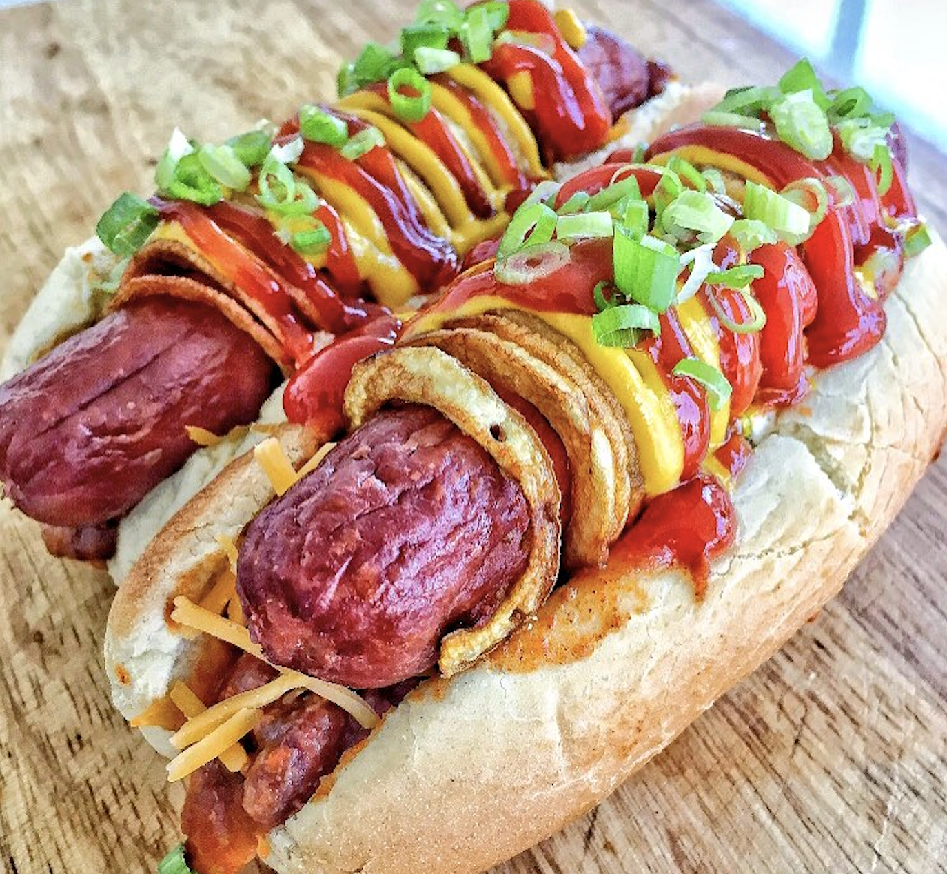 Two hot dogs in buns topped with crispy fried onion, ketchup, mustard, shredded cheese, and chopped green onions, served on a wooden surface.