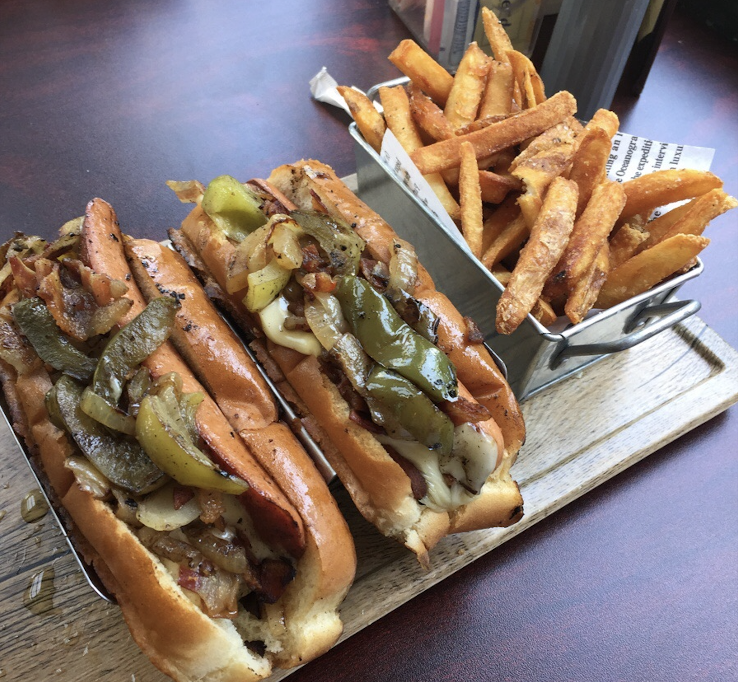 Two grilled hot dogs topped with peppers, onions, and cheese sit on a wooden tray beside a small basket of seasoned French fries.