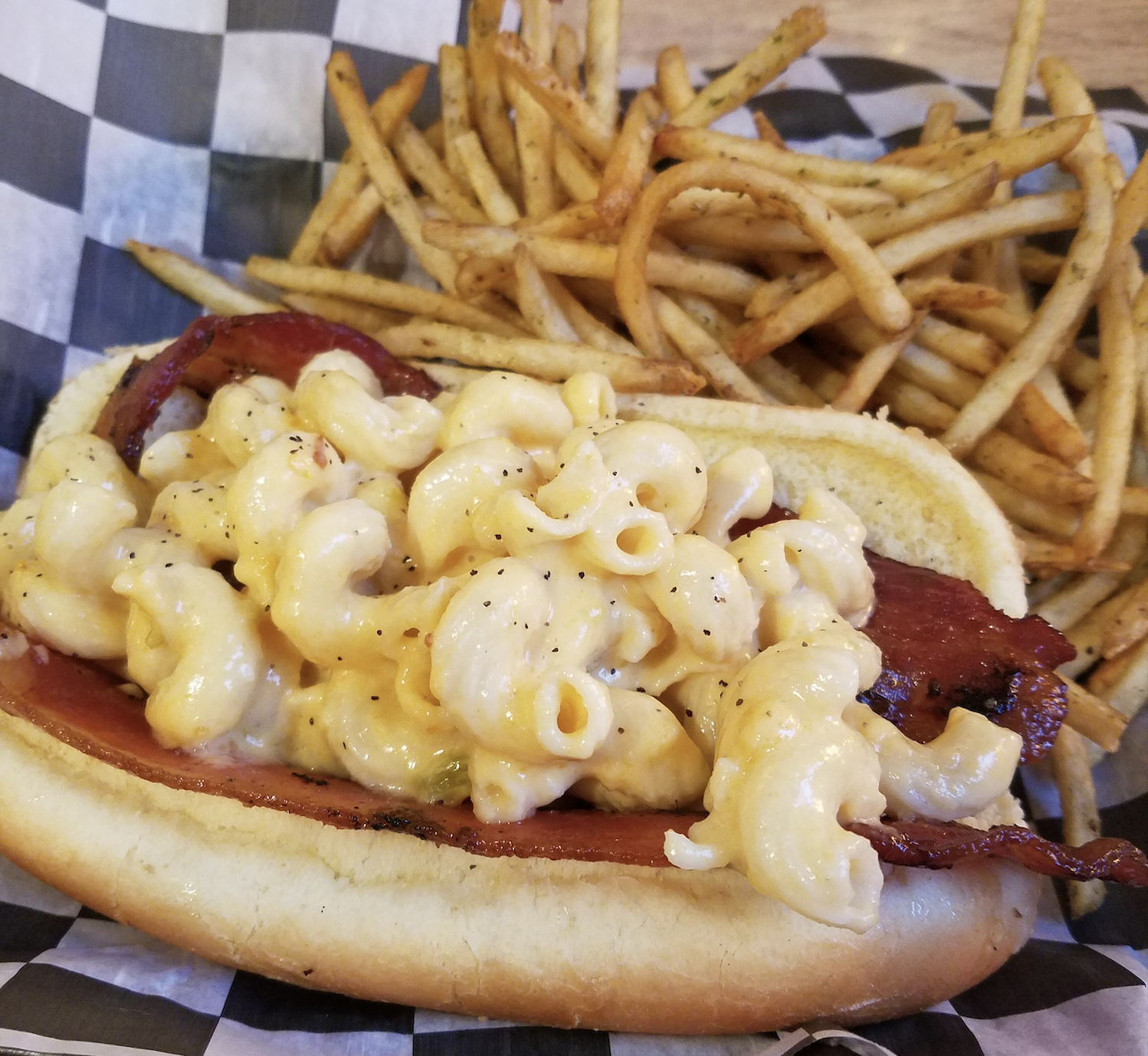 A hot dog bun filled with bacon and creamy macaroni and cheese, served with a side of thin, crispy French fries on a checkered paper.