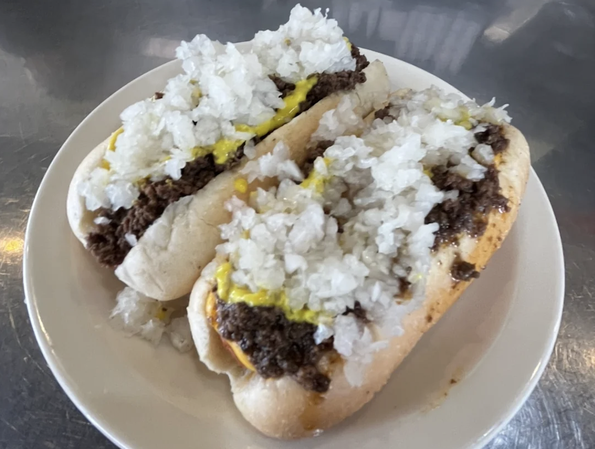 Two hot dogs on a white plate, topped with chopped onions, ground beef chili, and yellow mustard, on a shiny metal surface.