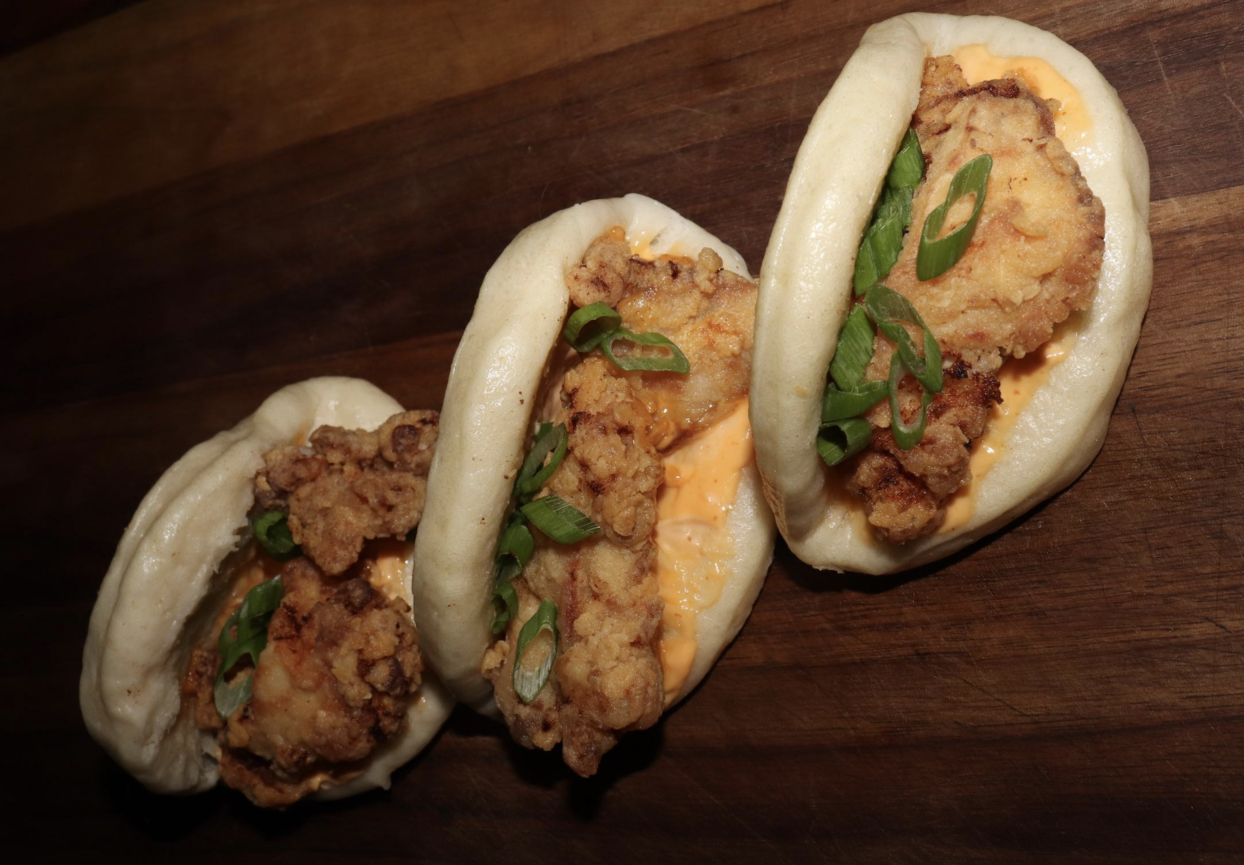 Three steamed bao buns filled with fried chicken, orange sauce, and sliced green onions are arranged on a wooden surface.