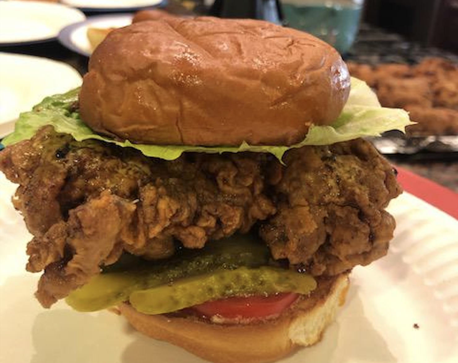 A fried chicken sandwich with lettuce, pickles, tomato, and a bun, served on a white plate.
