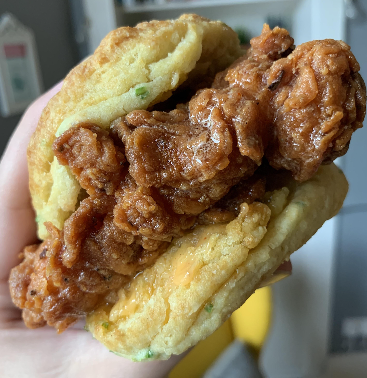 A hand holds a close-up of a fried chicken sandwich with crispy chicken and sauce between two fluffy biscuits.