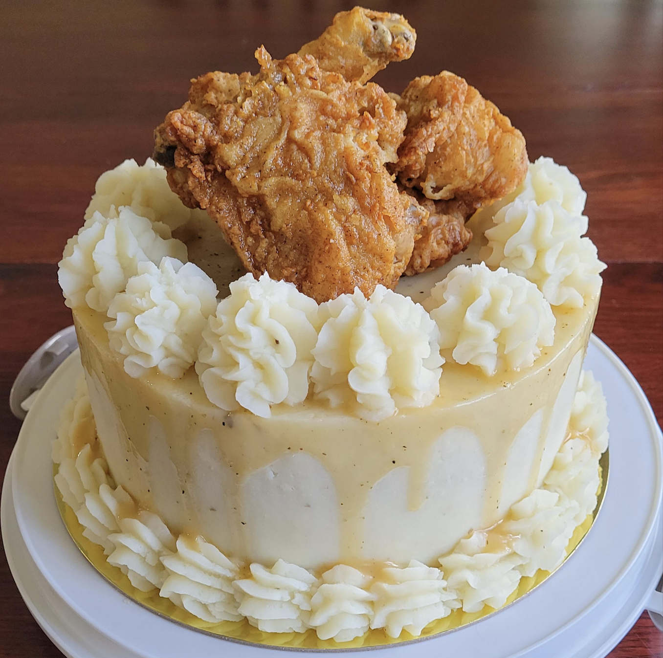 A round cake decorated with white frosting and topped with three pieces of fried chicken. The cake sits on a white plate and has piped frosting around the edges.