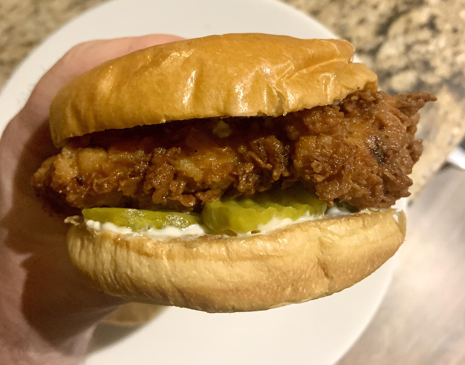 A hand holds a fried chicken sandwich with pickles and mayonnaise on a soft, golden bun. A white plate and brown countertop are in the background.