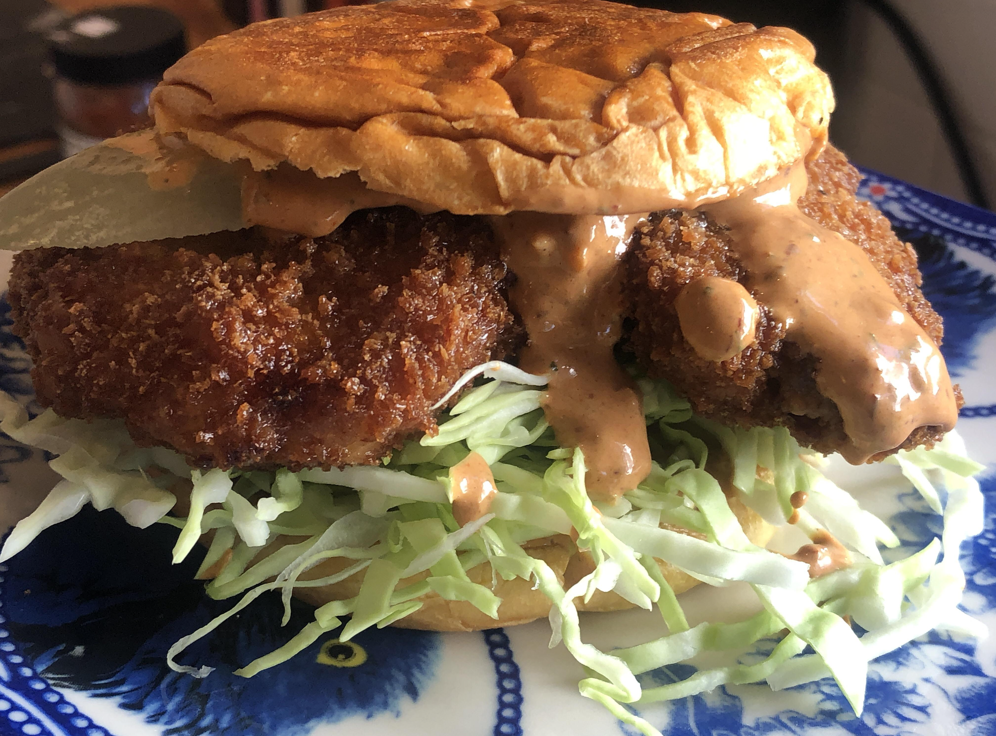 A crispy fried chicken sandwich with shredded lettuce, sauce, and a pickle on a blue-patterned plate.