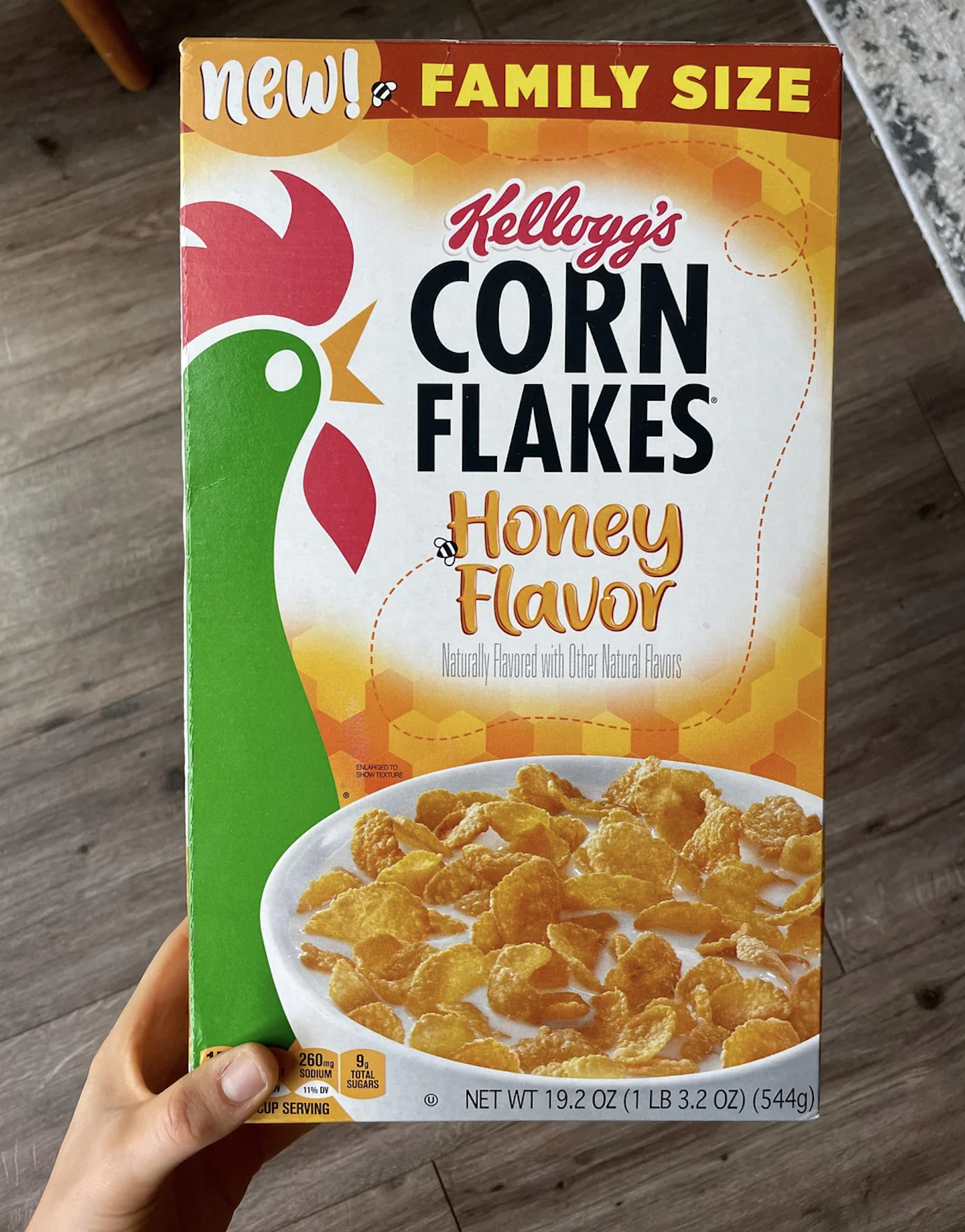 A hand holds a family-size box of Kellogg’s Corn Flakes Honey Flavor cereal, featuring a rooster, cereal bowl, and nutritional details on the front.