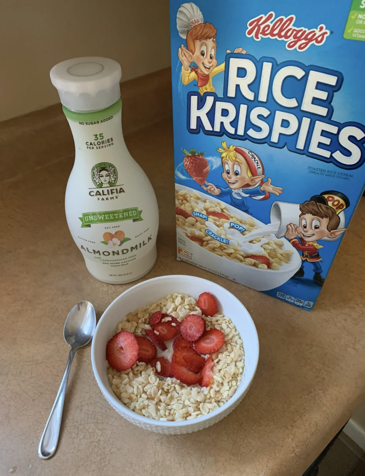 A bowl of Rice Krispies cereal with sliced strawberries and almond milk sits on a countertop, next to a spoon, a bottle of Califia Farms almond milk, and a box of Kellogg’s Rice Krispies.