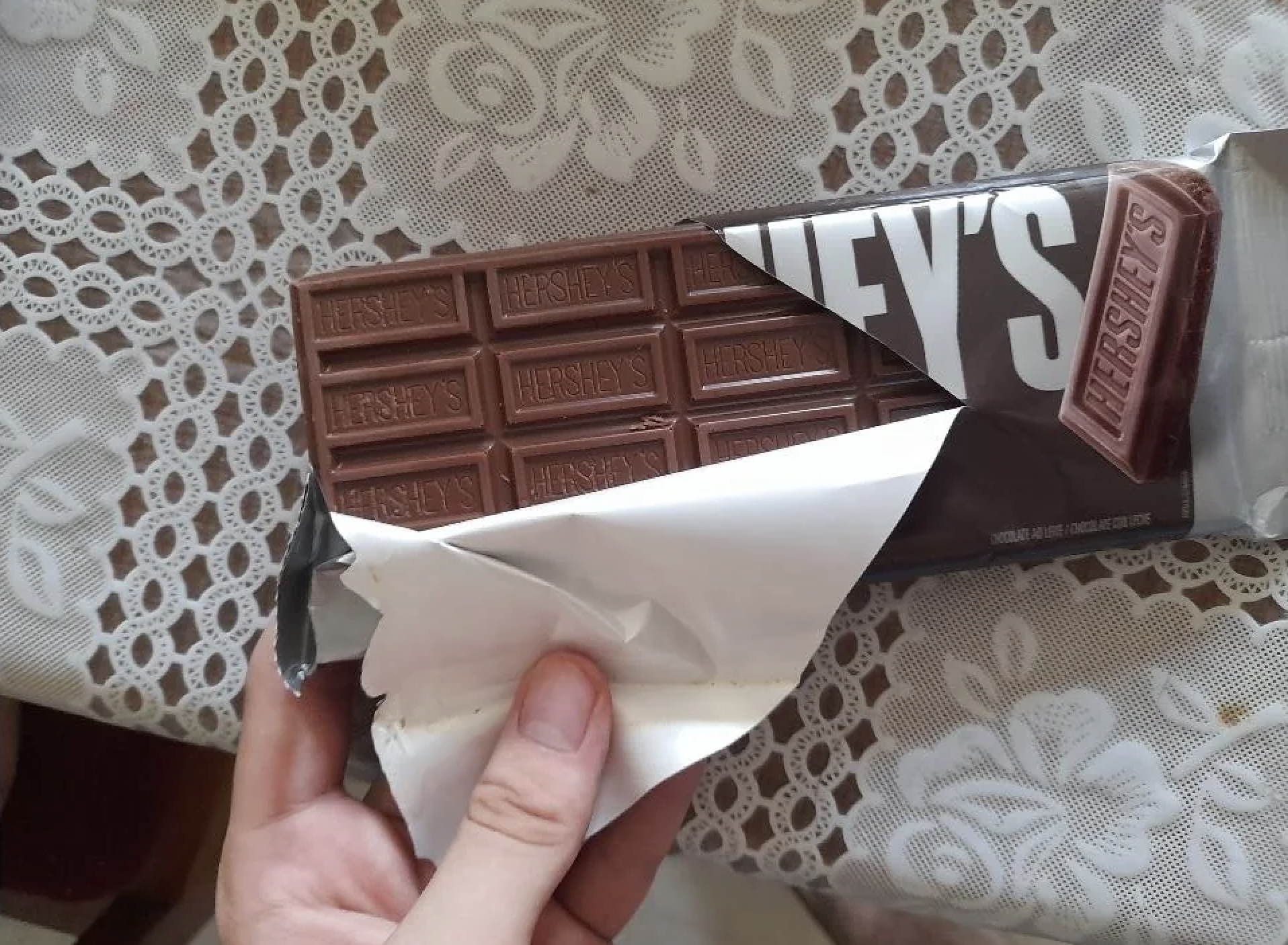 A hand holding a partially unwrapped Hershey’s chocolate bar on a table with a white lace tablecloth.