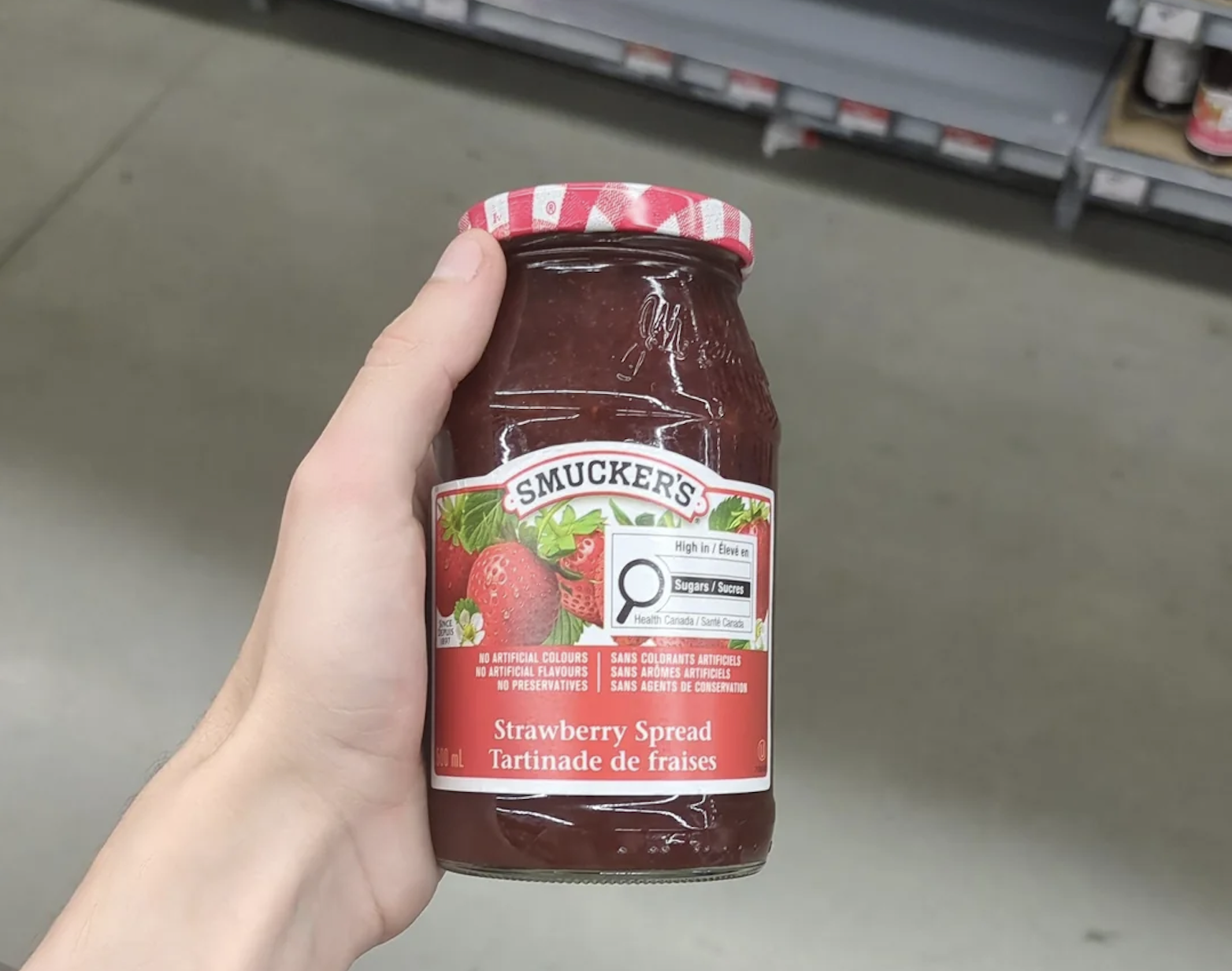 A hand holds a jar of Smucker’s Strawberry Spread in a store aisle. The label shows strawberries and text in English and French. Shelves are visible in the background.
