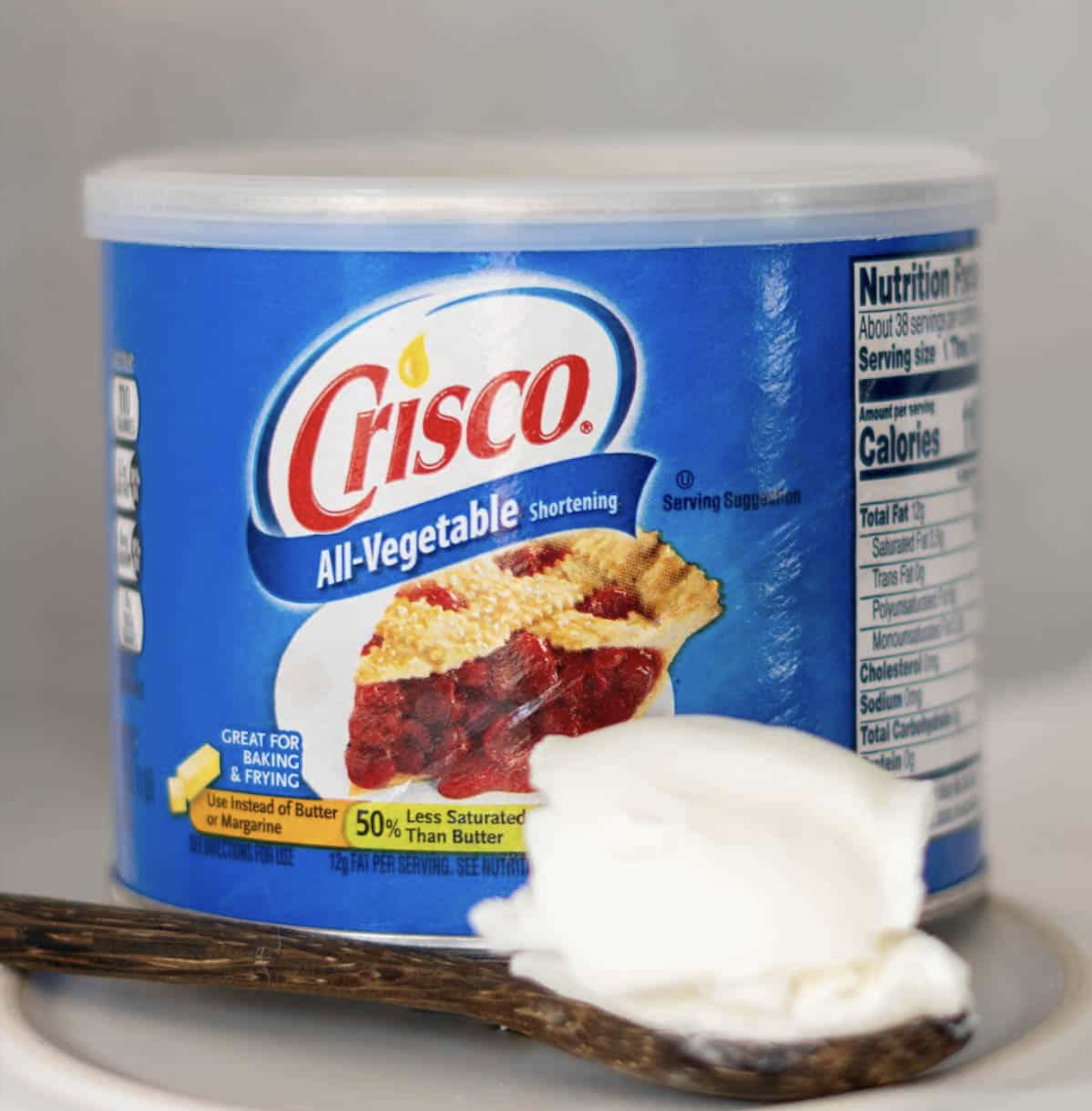 A container of Crisco All-Vegetable Shortening with a slice of pie on the label sits next to a spoonful of white shortening. Nutrition facts are partially visible in the background.