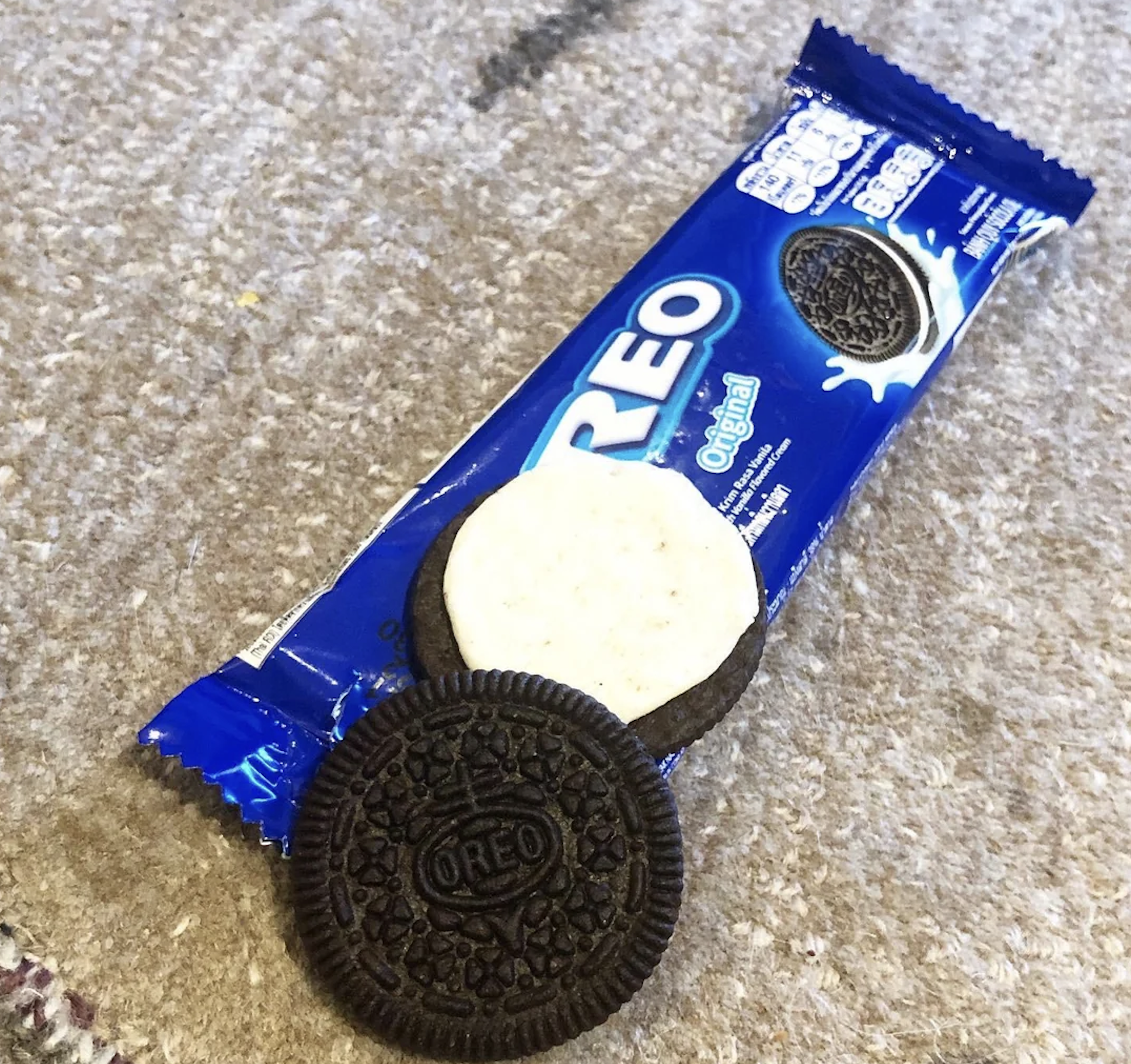 An opened Oreo cookie with its cream exposed sits on top of a blue Oreo Original package, placed on a beige carpet.