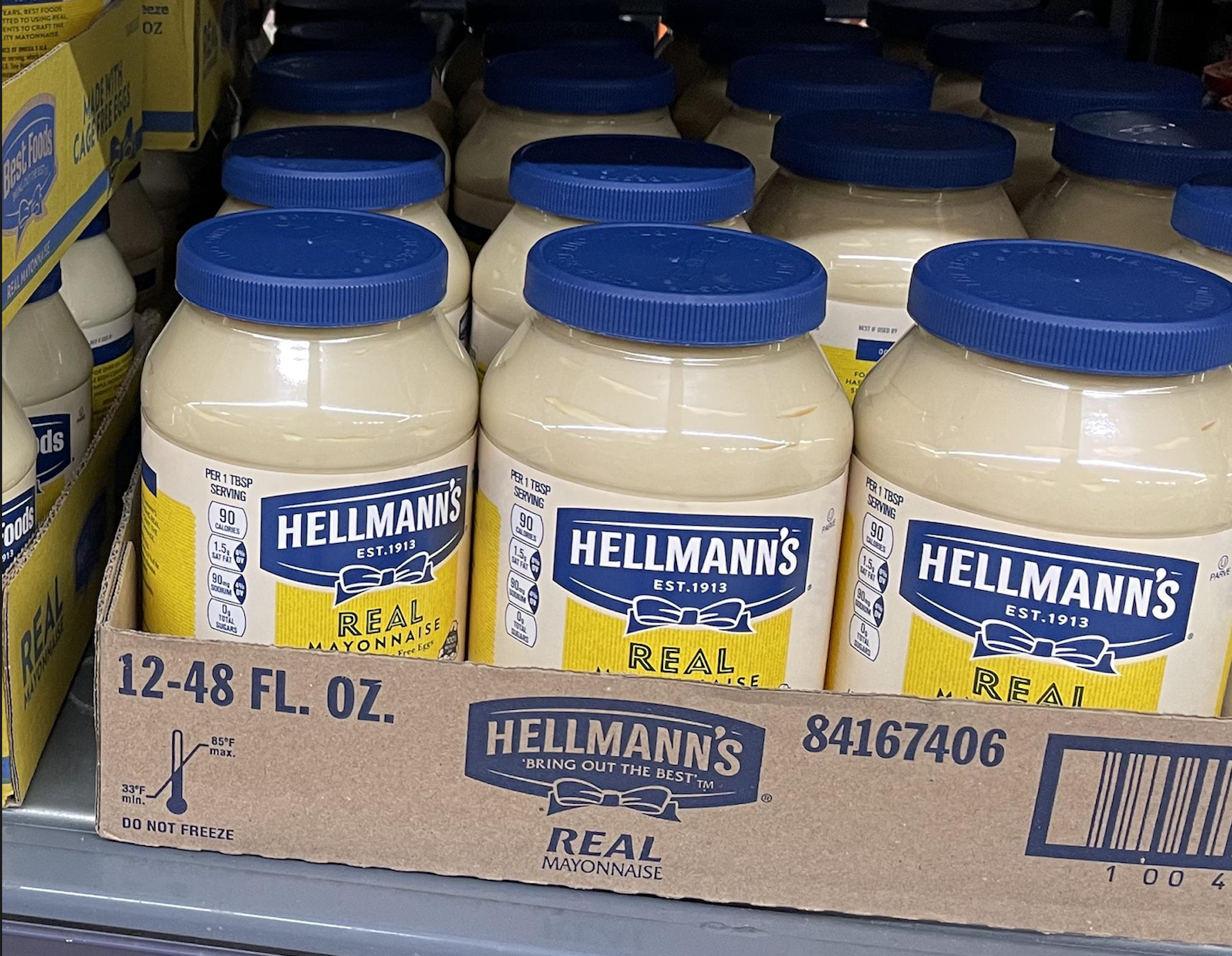 Several large jars of Hellmann’s Real Mayonnaise are displayed in a cardboard box on a store shelf. The jars have blue lids and yellow labels.