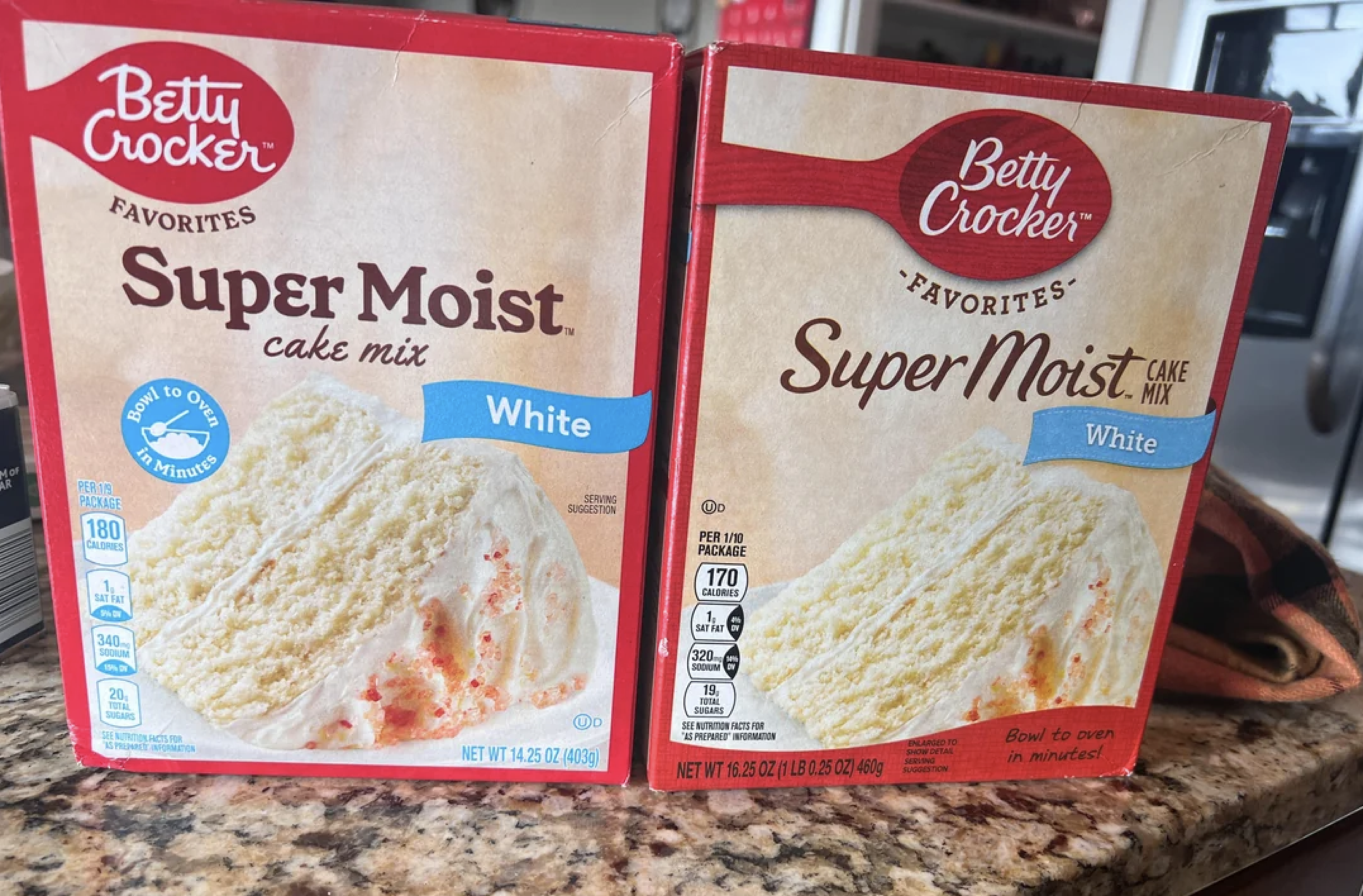 Two boxes of Betty Crocker Super Moist White Cake Mix sit side by side on a kitchen counter. Both boxes show a slice of white cake with frosting on the front.