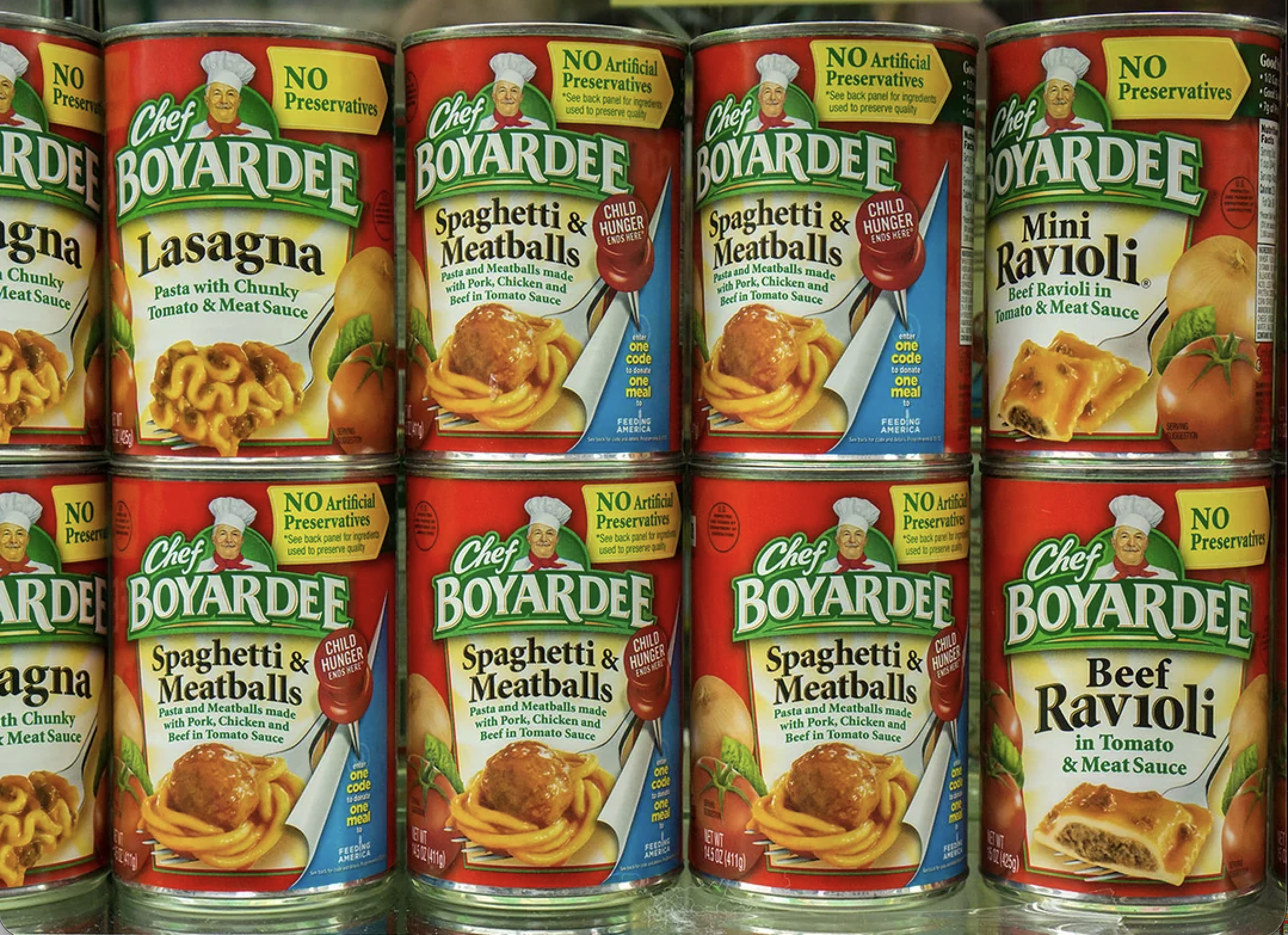Cans of Chef Boyardee pasta including lasagna, spaghetti & meatballs, mini ravioli, and beef ravioli, stacked on a supermarket shelf. Labels say “No Artificial Preservatives.”