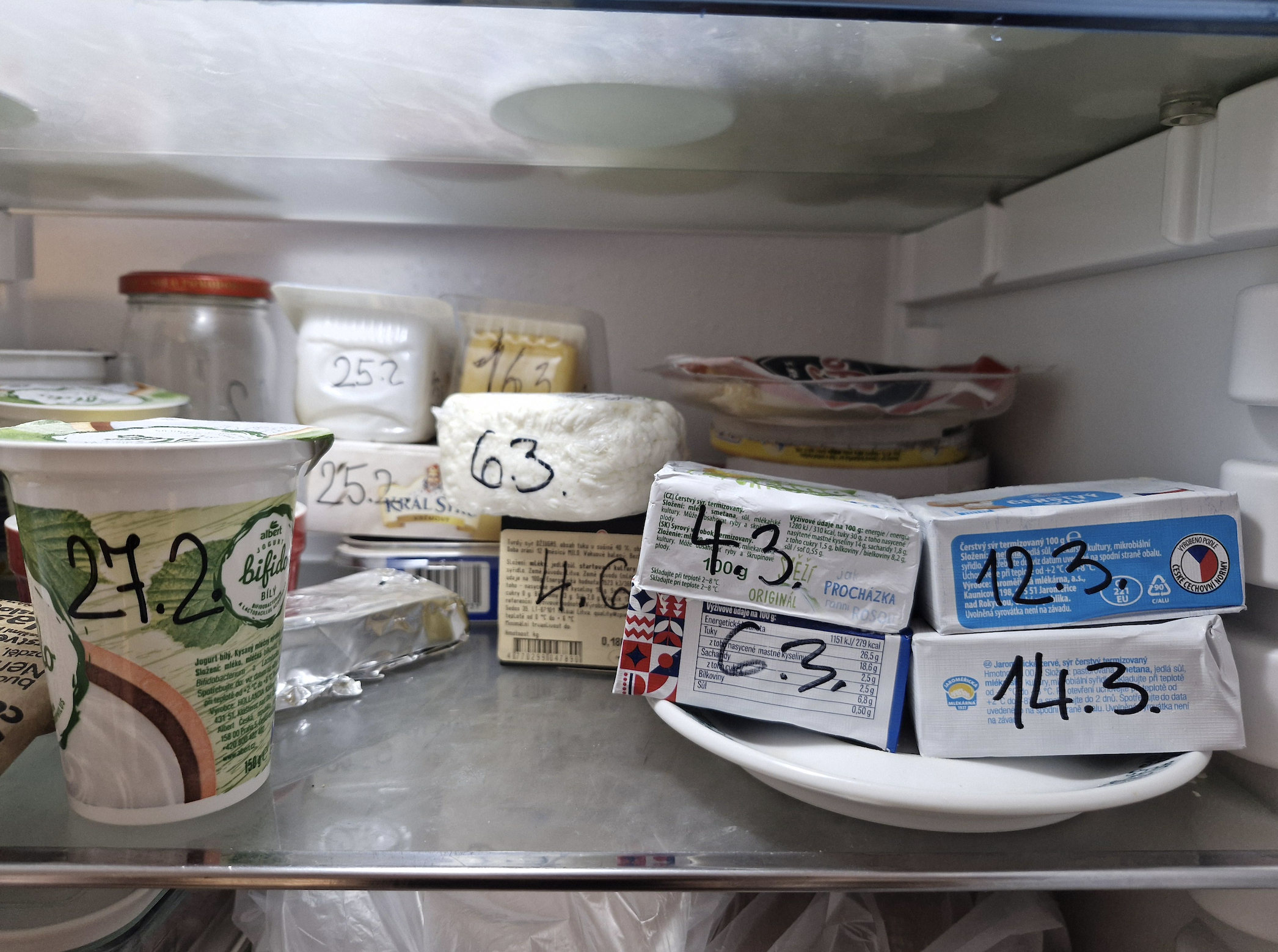 An open fridge shelf with various dairy products labeled with handwritten dates, including yogurt cups, blocks of cheese, and jars, all marked with black marker for expiration or opening dates.