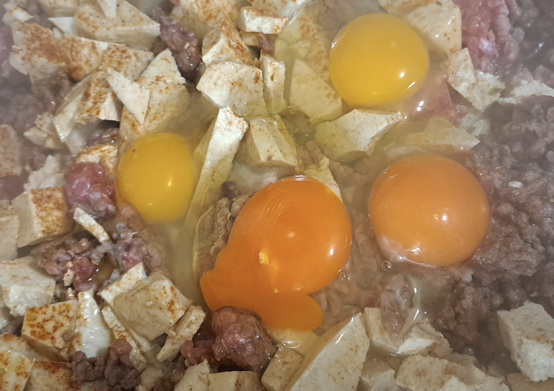 Raw eggs cracked over a mixture of diced tofu, ground meat, and seasonings, ready to be cooked together in a pan.
