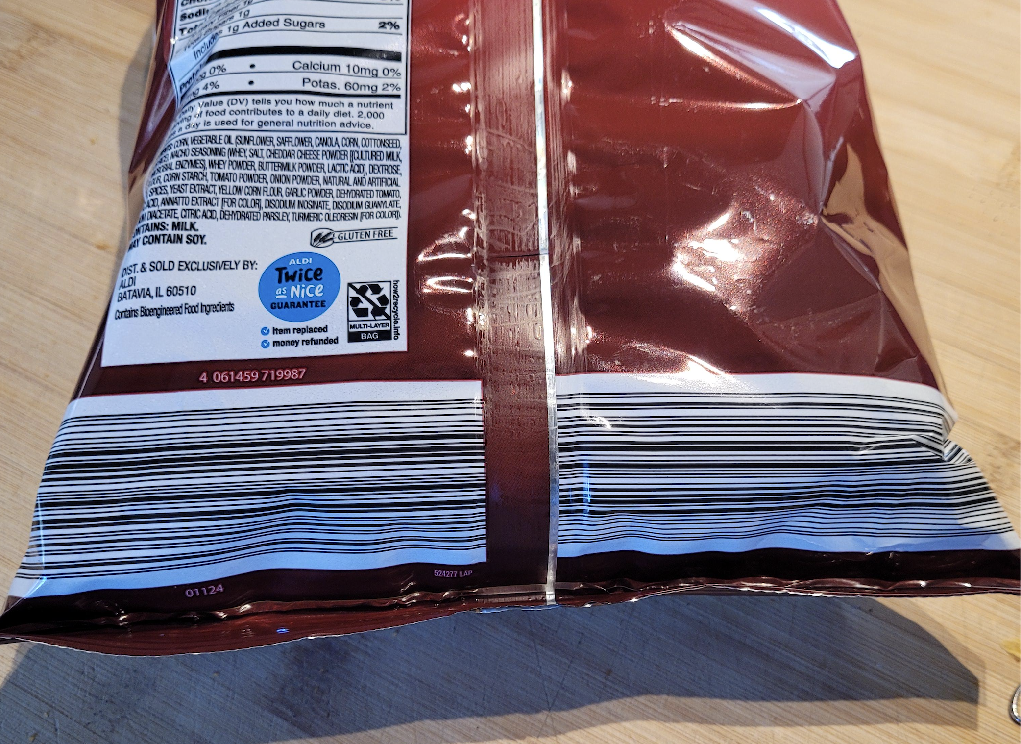 A close-up of the back of a maroon snack bag, showing a barcode, nutrition label, and recycling information on a wooden surface.