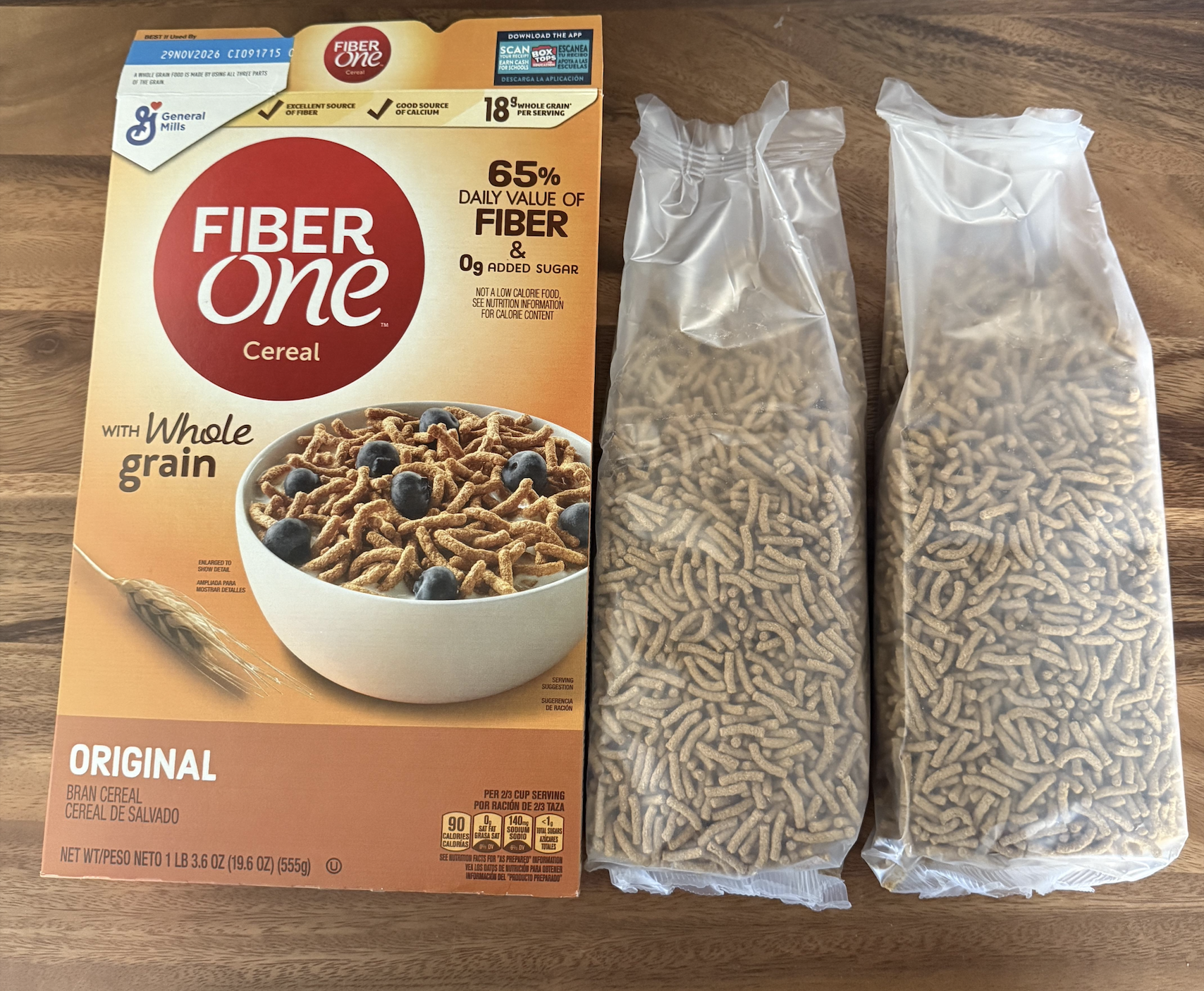 A box of Fiber One Original cereal is shown on a wooden surface, with two clear plastic bags filled with bran cereal sticks placed next to it.