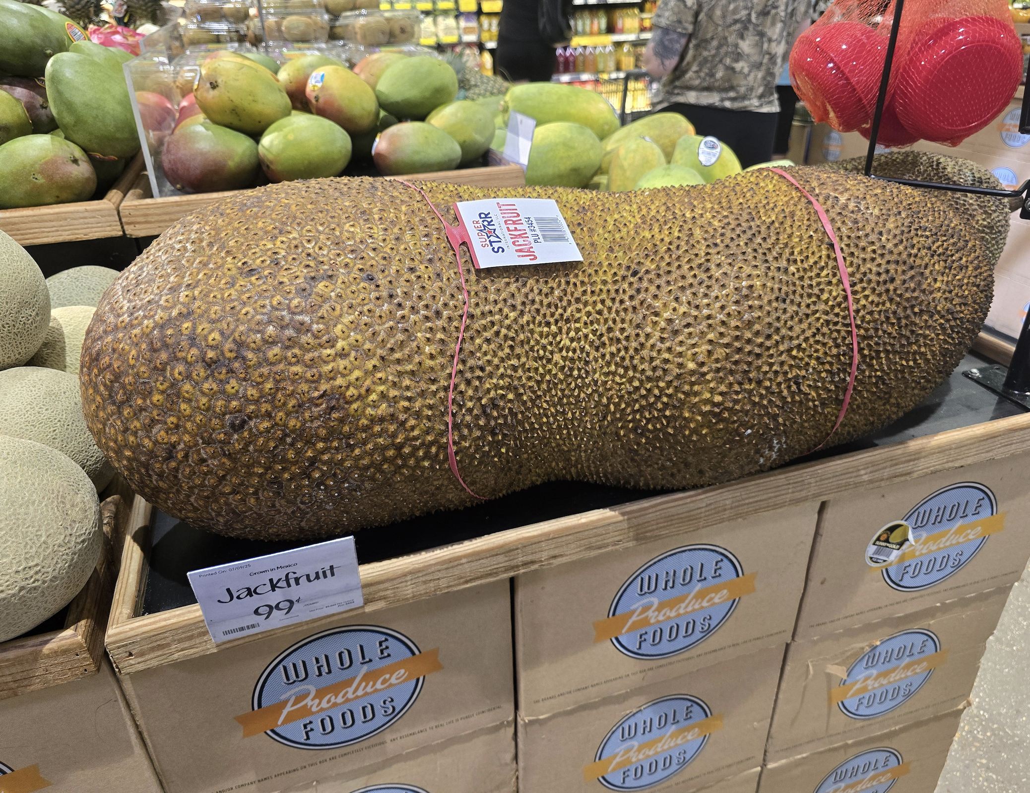 A large jackfruit with rough, spiky skin rests on boxes at a Whole Foods store. Nearby are melons and mangos. A sign in front labels it as “Jackfruit” and shows the price per pound.